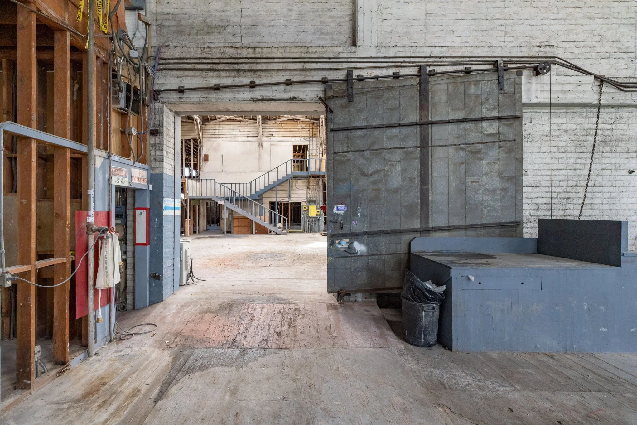 Interior of an industrial or warehouse space with exposed brick walls, electrical wiring, a metal sliding door, and a staircase leading upstairs.