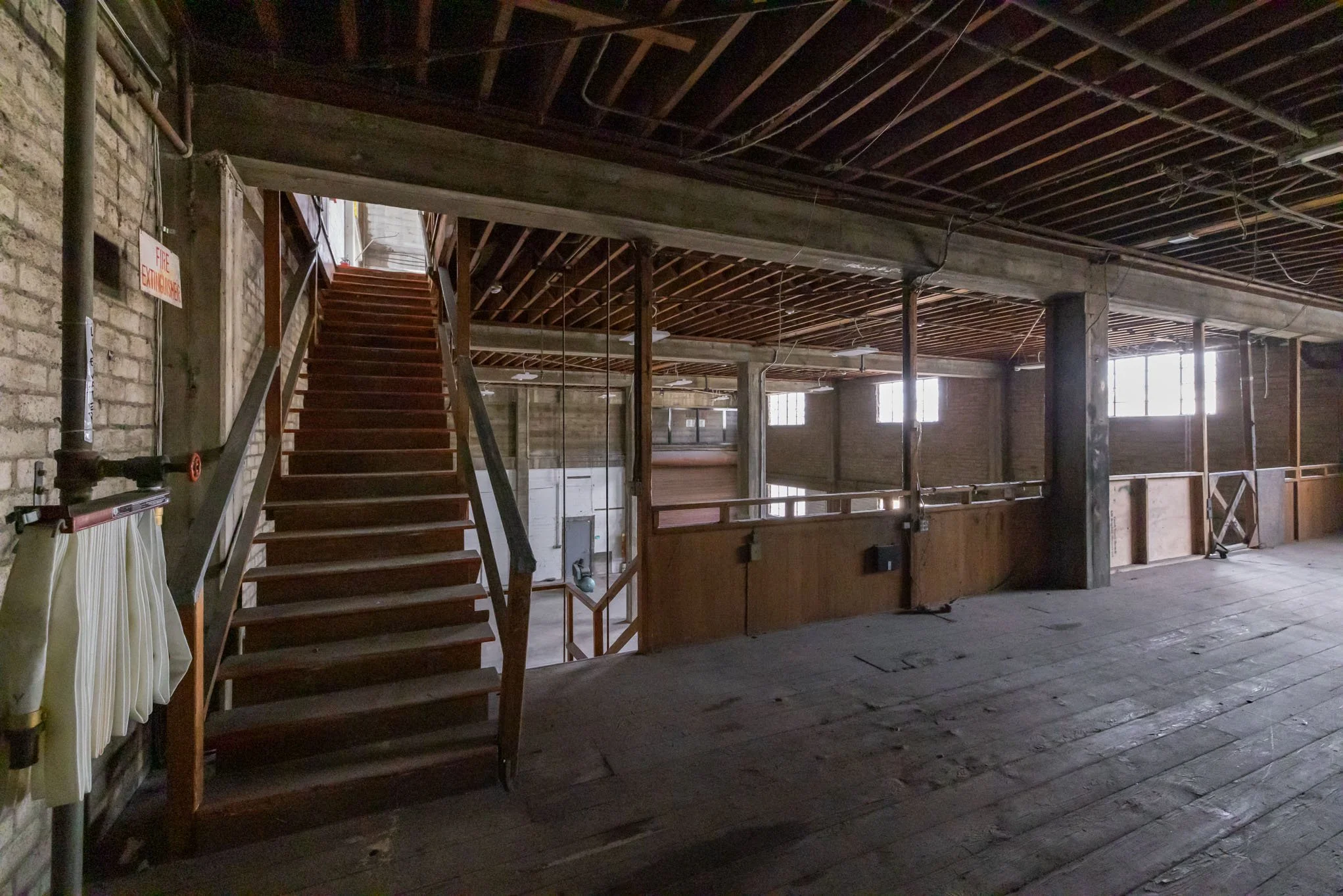 Interior view of an unfinished building with exposed brick walls, wooden flooring, a staircase leading to an upper level, and large windows allowing natural light.