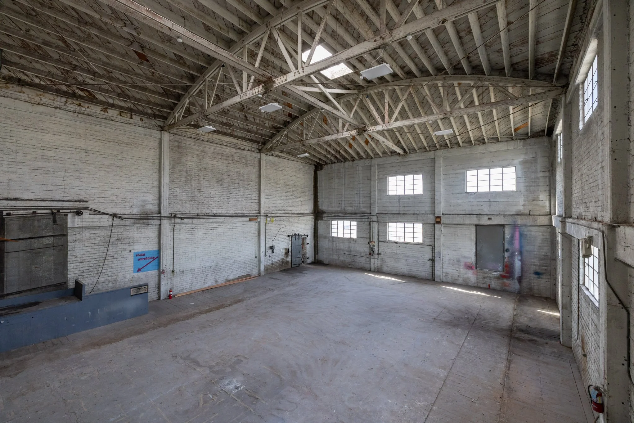 Empty warehouse with high ceiling, exposed wooden beams, concrete walls, and large windows providing natural light.