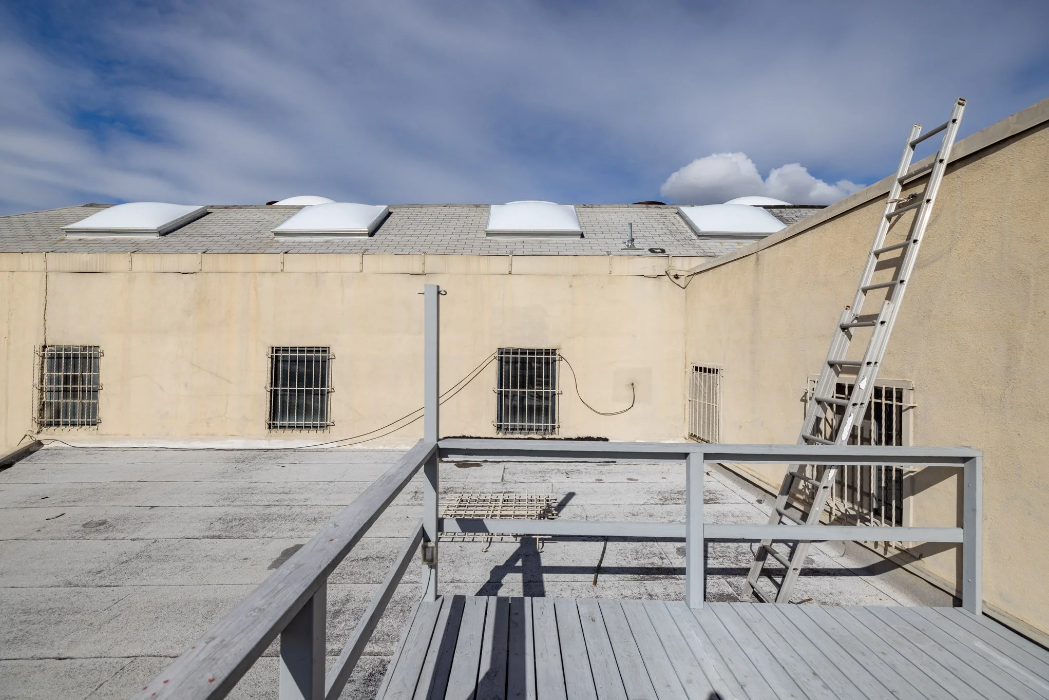View of a rooftop with skylights, metal bars on windows, a ladder leaning against a wall, and a wooden balcony railing under a partly cloudy sky.