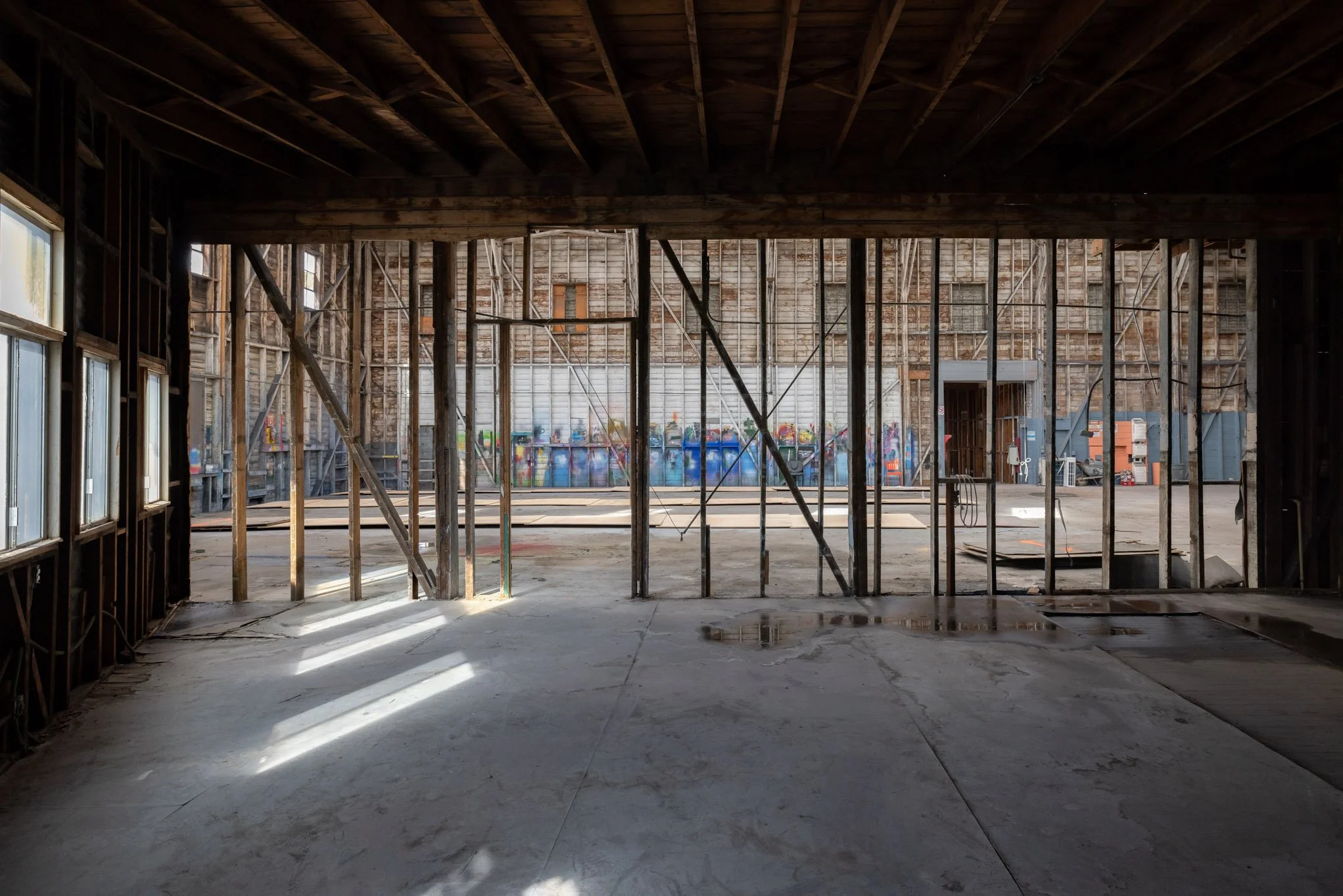 Interior of a building under renovation with exposed wooden and metal framing, sunlight streaming through windows, and graffiti on the far wall.