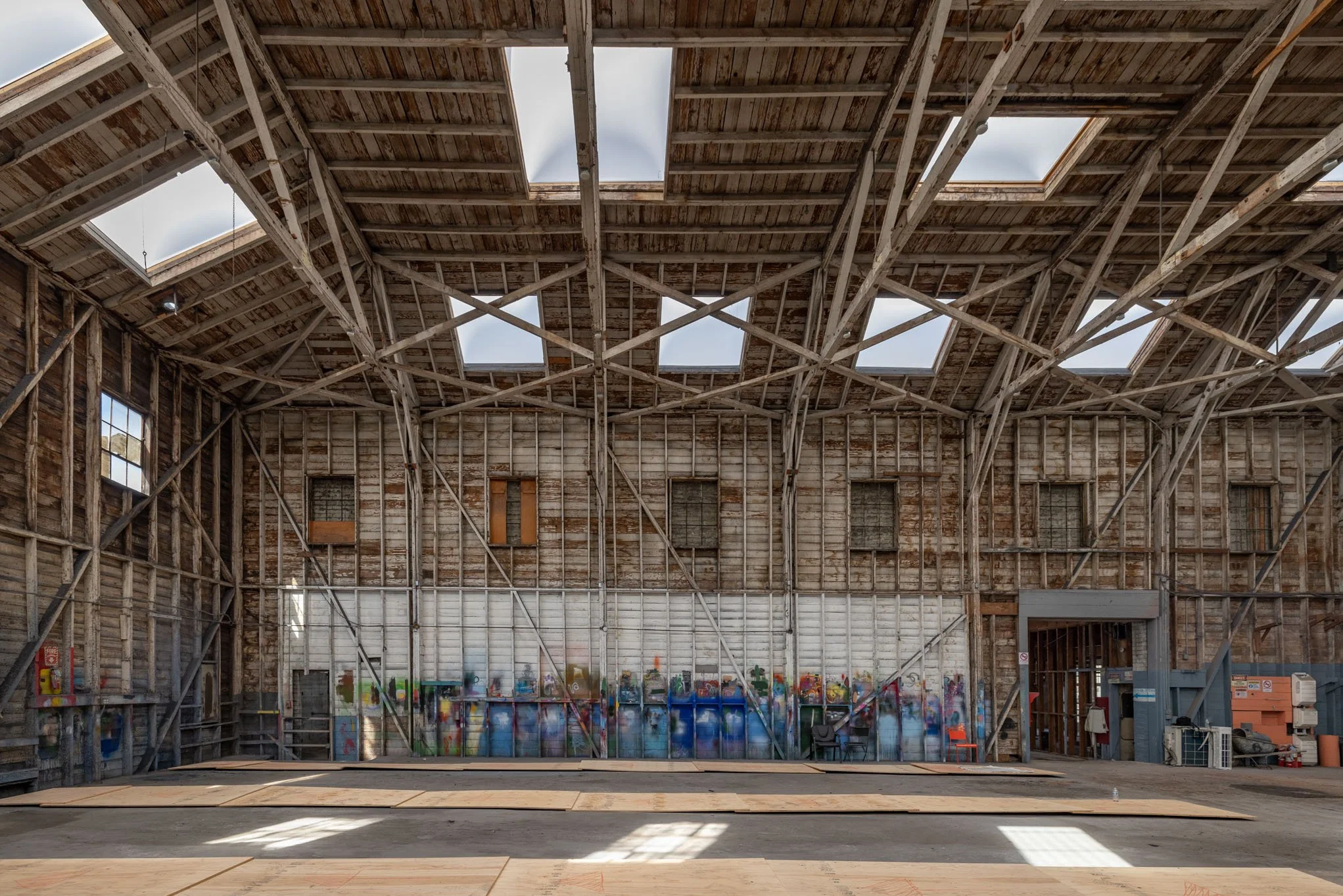 Inside an unfinished wooden building, exposed beams and walls, with multiple skylights in the roof allowing natural light, and a partially painted graffiti mural on the back wall.