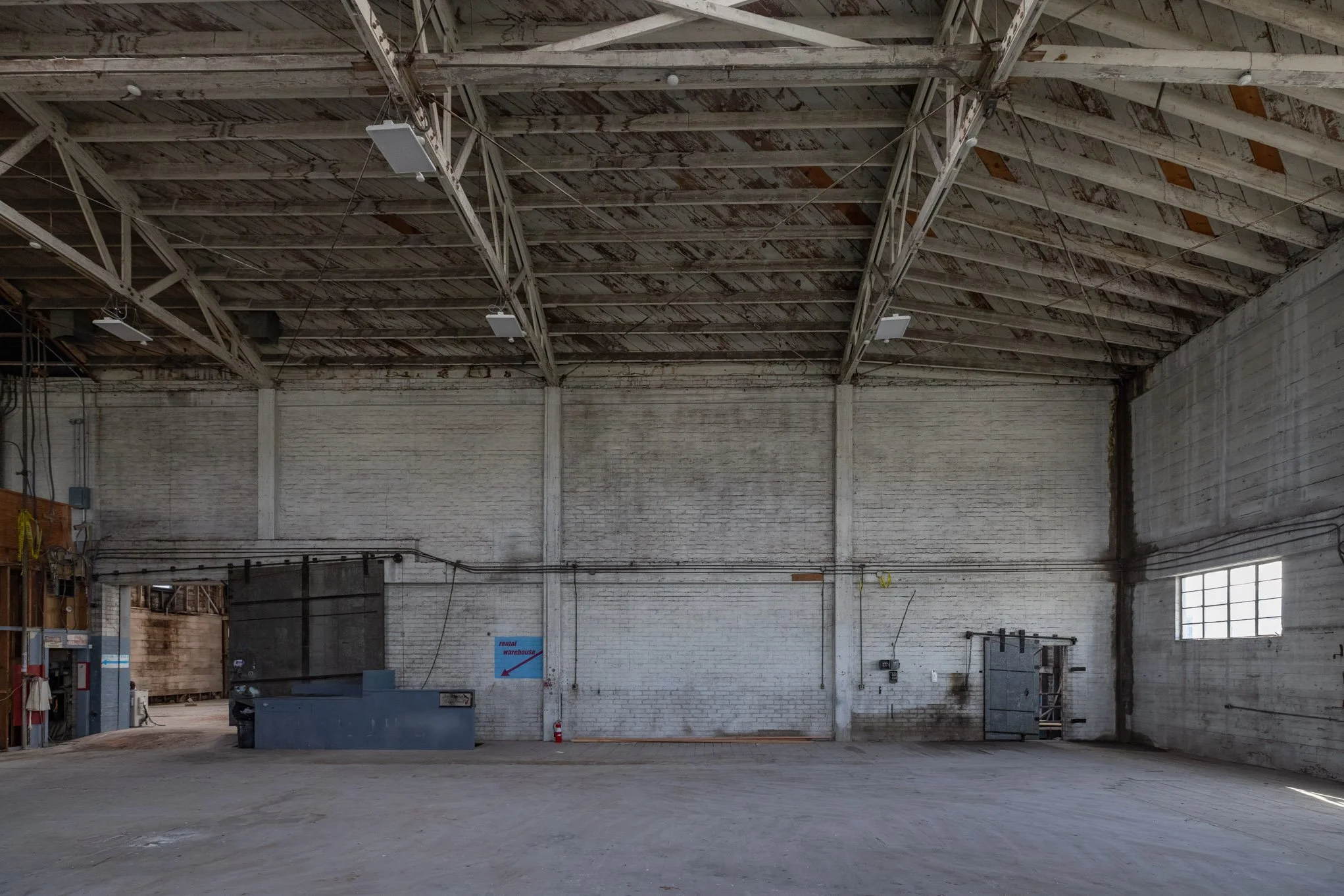 Empty industrial warehouse with white brick walls, high ceiling with exposed beams, and a large window on the right wall.