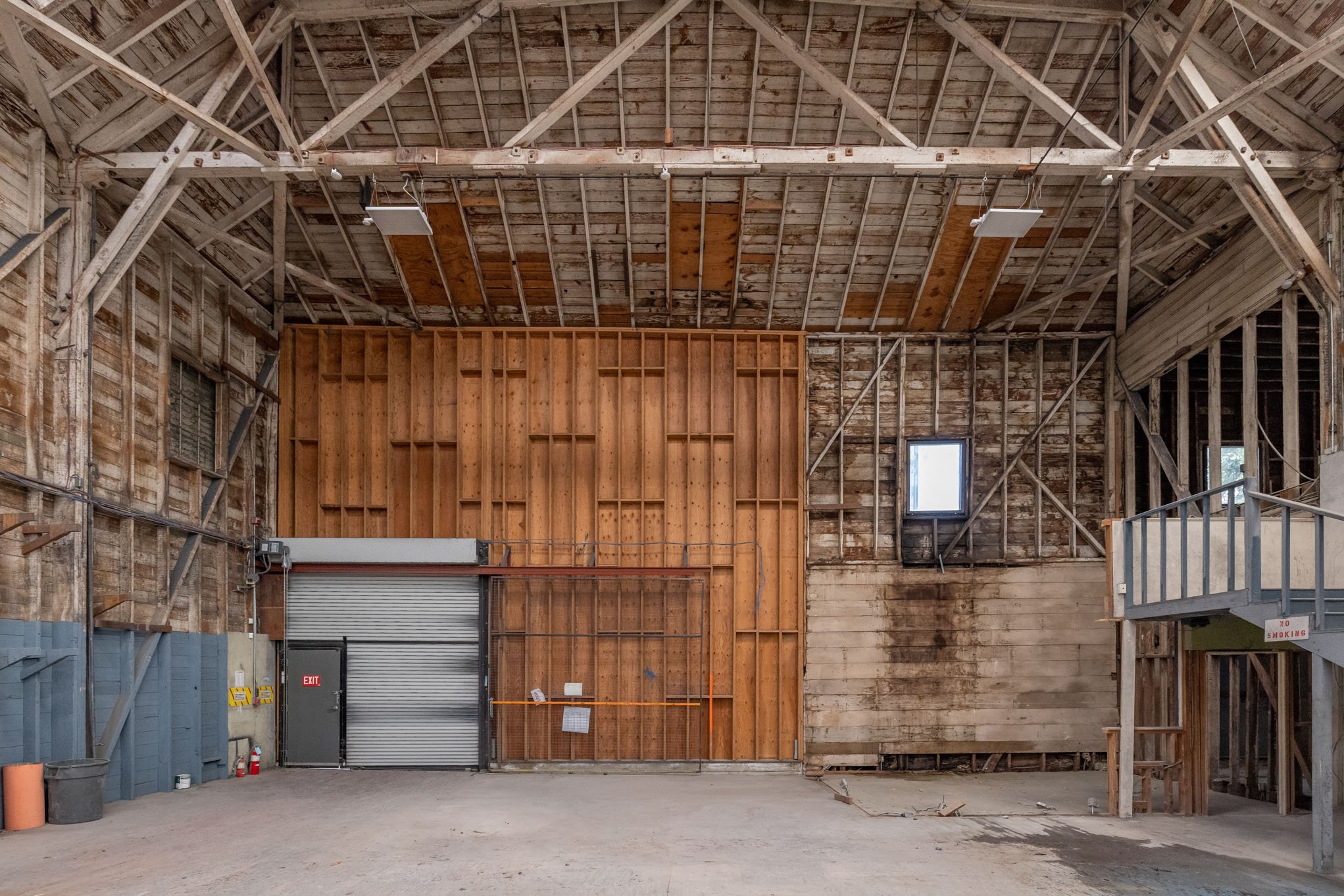 Interior of a large, unfinished warehouse or barn with exposed wooden framing, a metal roll-up door, small windows, and construction materials on the ground.