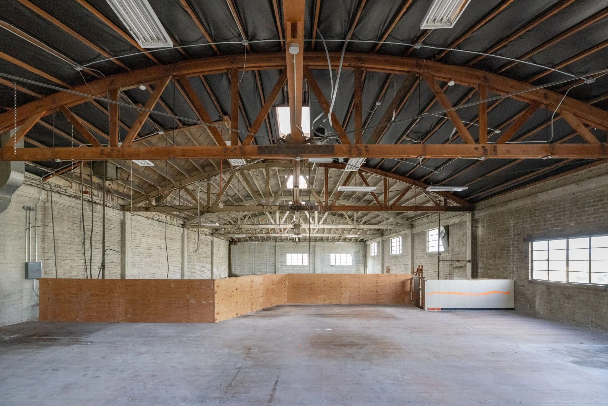 Empty industrial warehouse with exposed brick walls, large windows, and a high ceiling with wooden beams. A wooden barrier encloses a section near the back, and construction materials are visible.