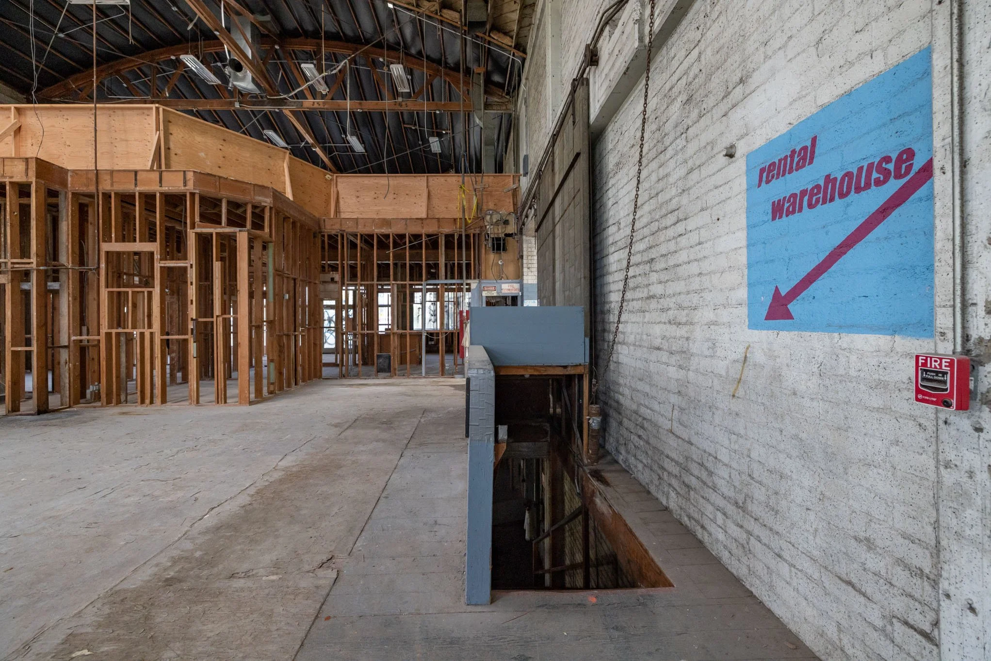 Interior of an unfinished rental warehouse with exposed wooden framing, a brick wall with a blue sign pointing downward, and a fire alarm box on the wall.