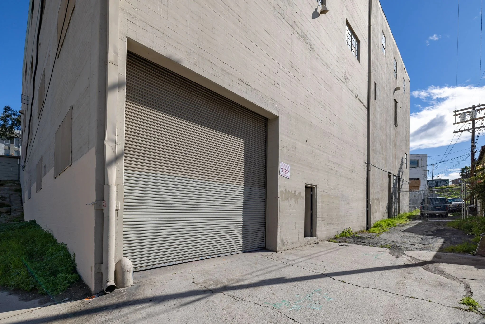 Concrete building with a large metallic rolling door, a small door, and a sign on the wall. Fenced alleyway and parked cars in the background.