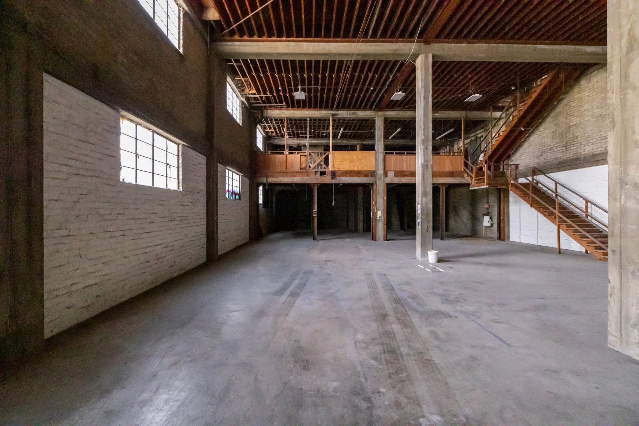 Interior of an unfinished industrial loft with exposed brick walls, large windows, a second-floor wooden balcony, and a staircase on the right side.