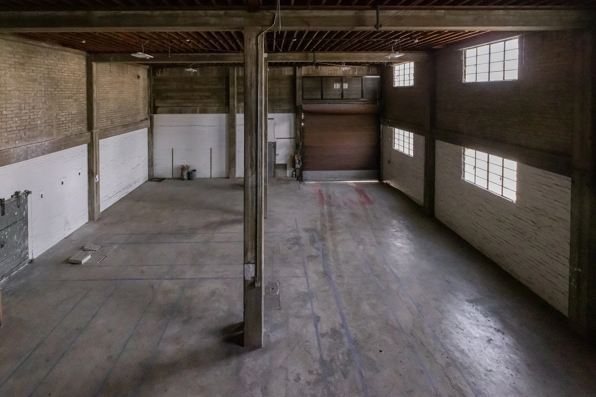 Empty industrial warehouse with brick and wood walls, concrete floor, large windows, and a rolling door.