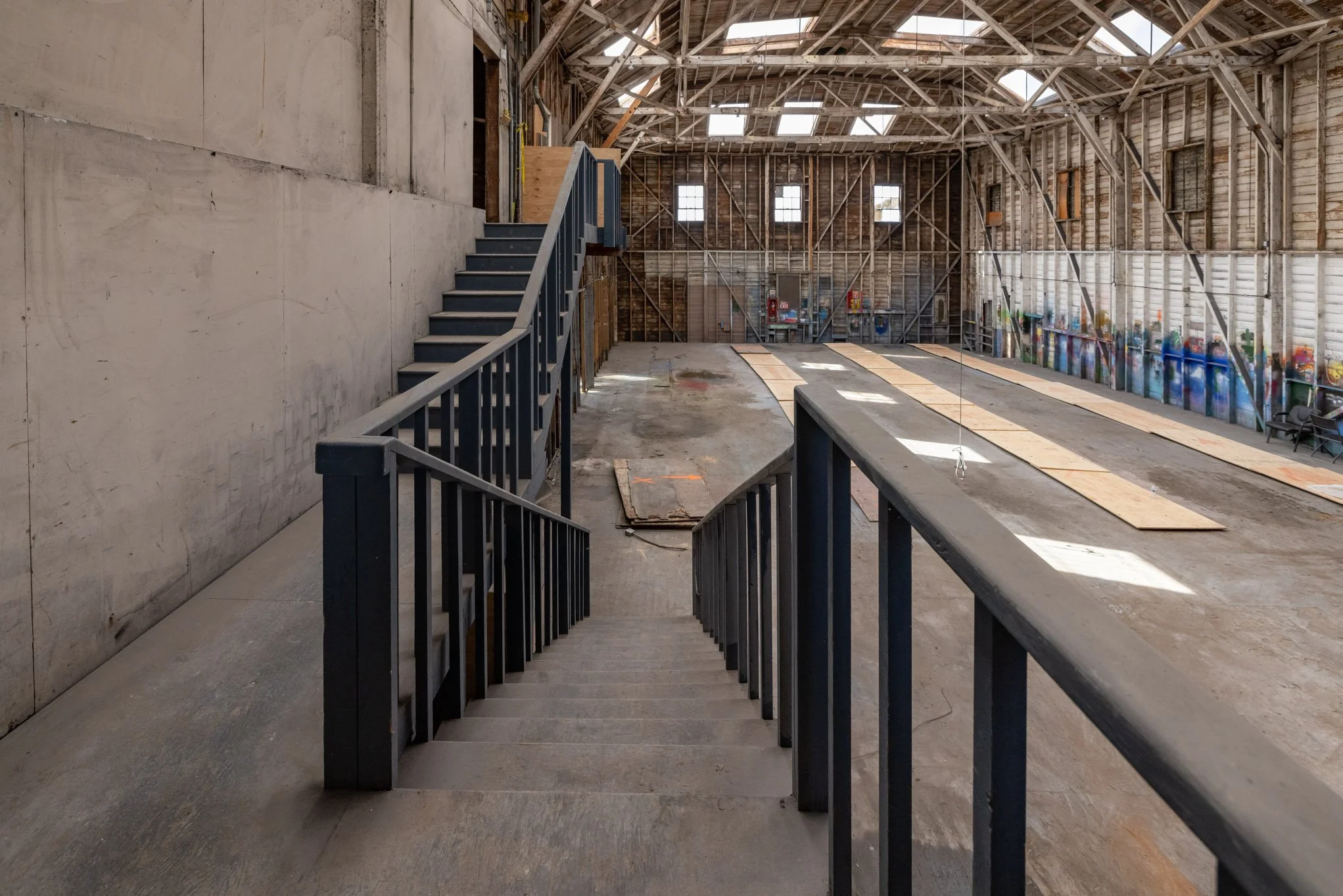 Interior of an unfinished industrial building with exposed wooden and metal framing, concrete flooring, a staircase, and graffiti on the walls.