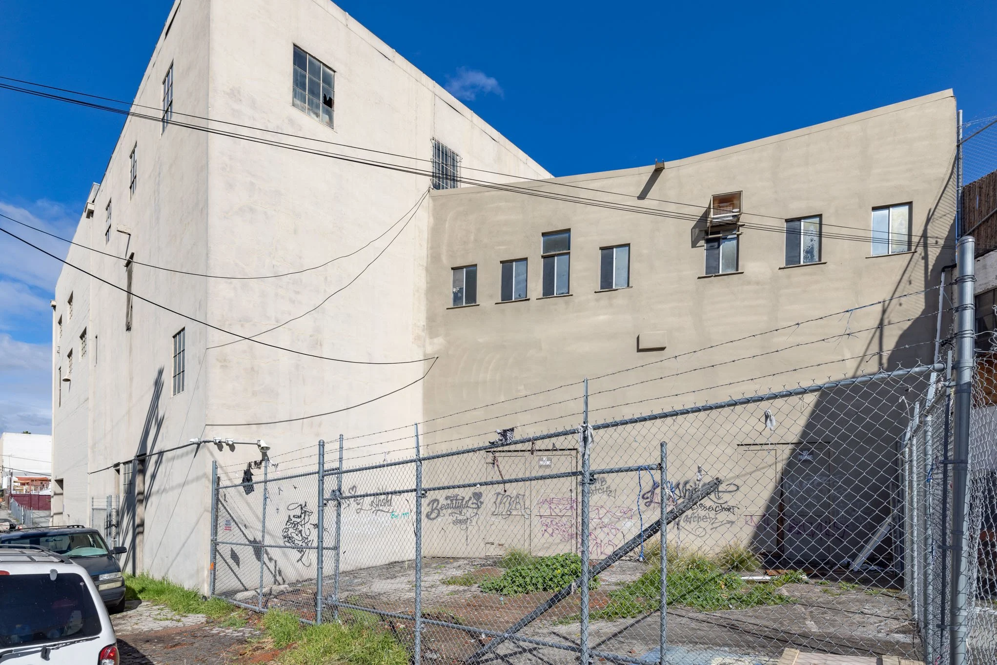A multi-story building with a white and beige exterior, multiple small windows, and scattered graffiti on a lower wall, surrounded by a chain-link fence with some overgrown weeds at the base.