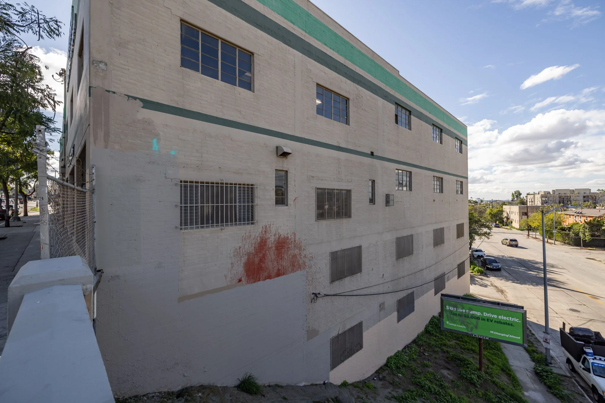 Side of a white brick building with barred and boarded-up windows, a small lower window with a security grill, some red paint splatter, and a green billboard promoting EV rebates. The street with parked cars and a partly cloudy sky in the background.