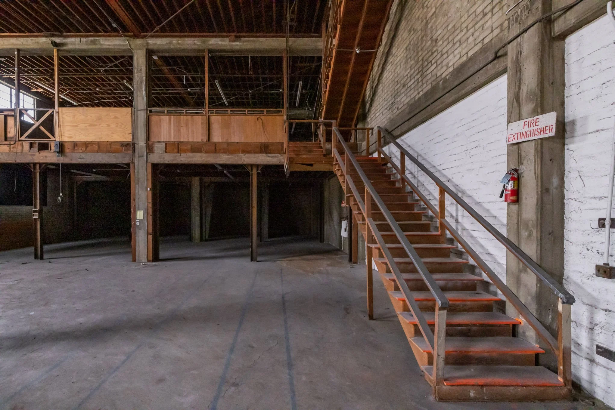 Empty industrial warehouse with wooden staircase leading to upper level, brick and concrete walls, and fire extinguisher mounted near staircase.