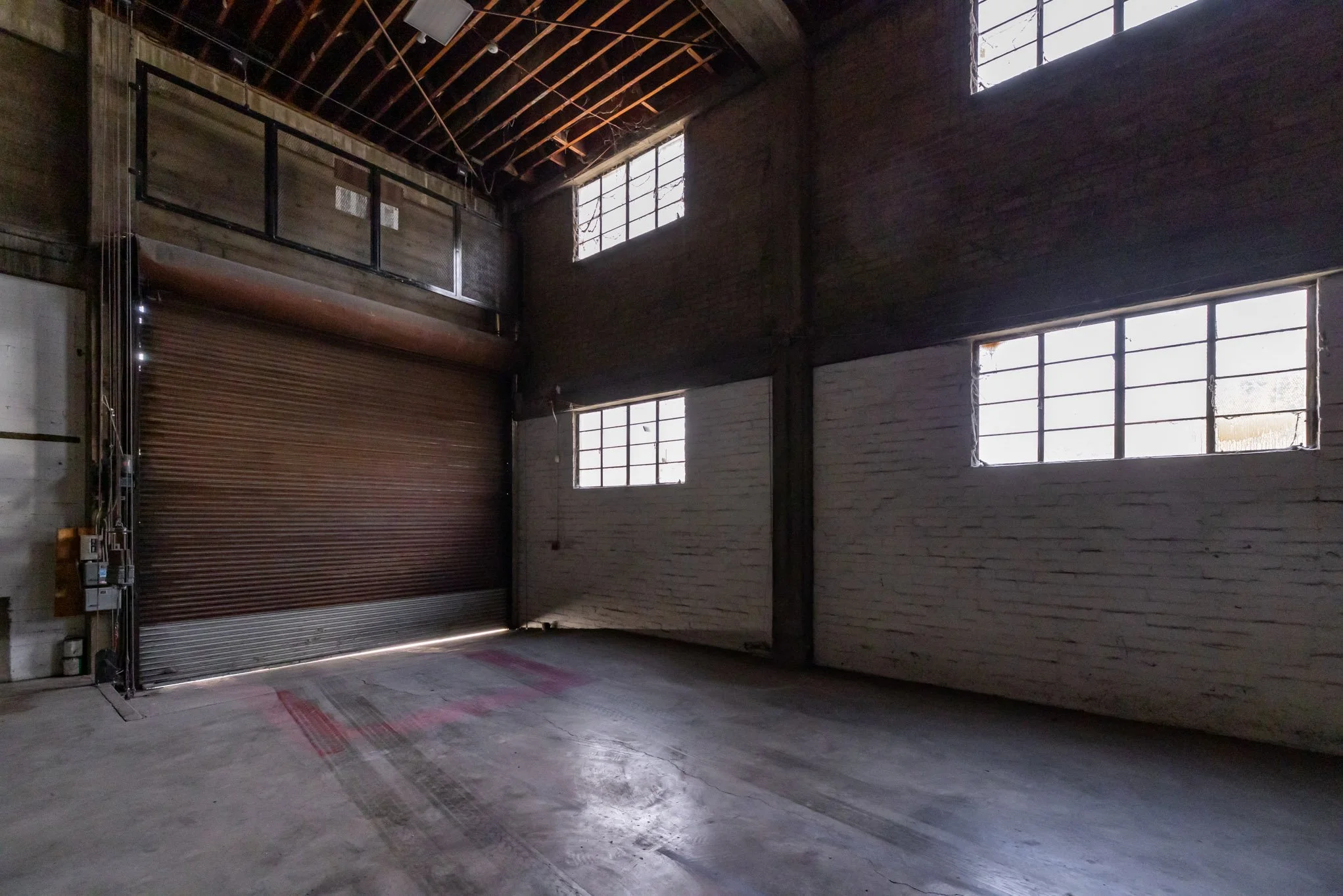Empty industrial warehouse with large metal roll-up door, exposed brick and concrete walls, and multiple high windows allowing natural light.