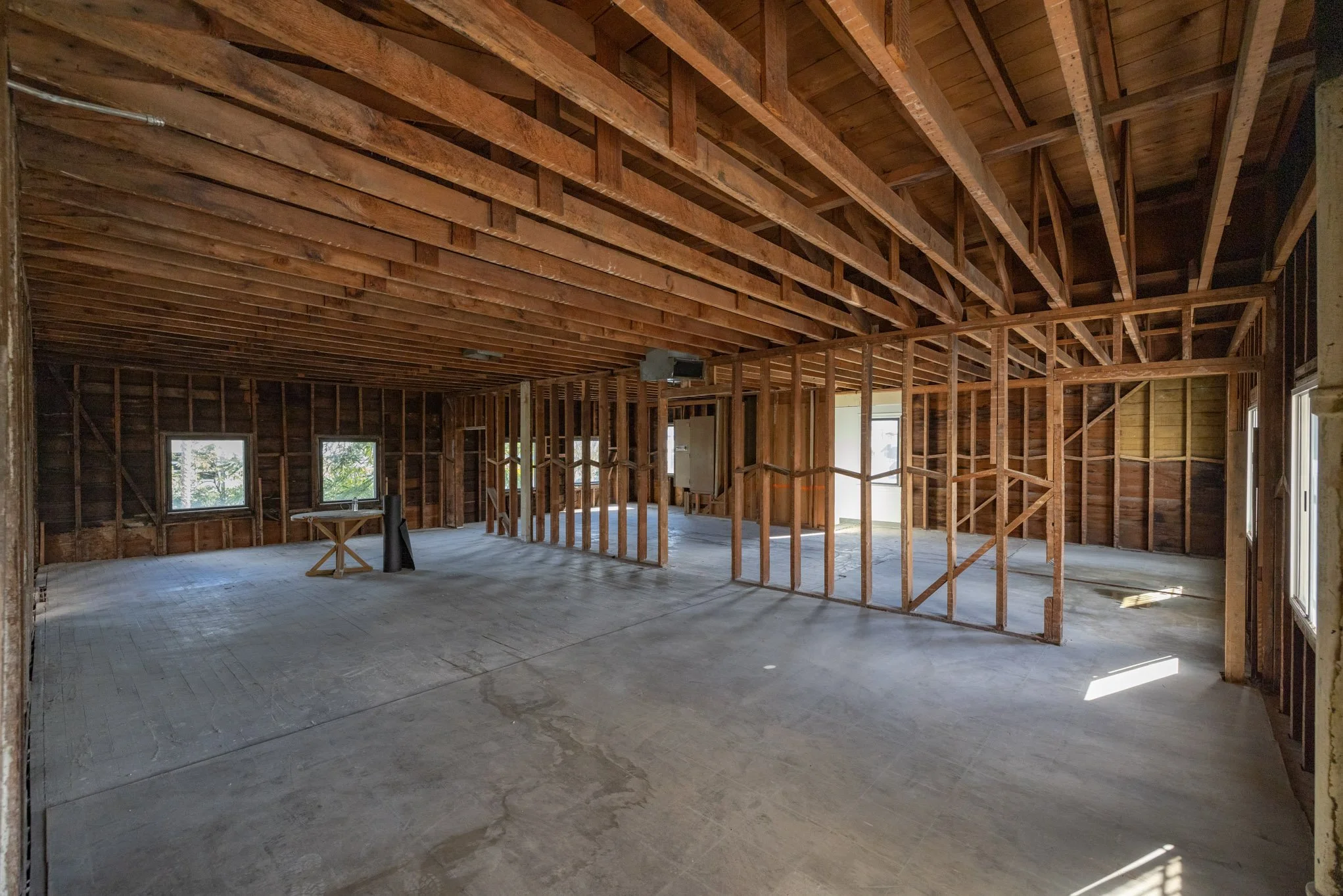 Interior of a house under construction with exposed wooden framing, ceiling beams, and a concrete floor, with several windows providing natural light.