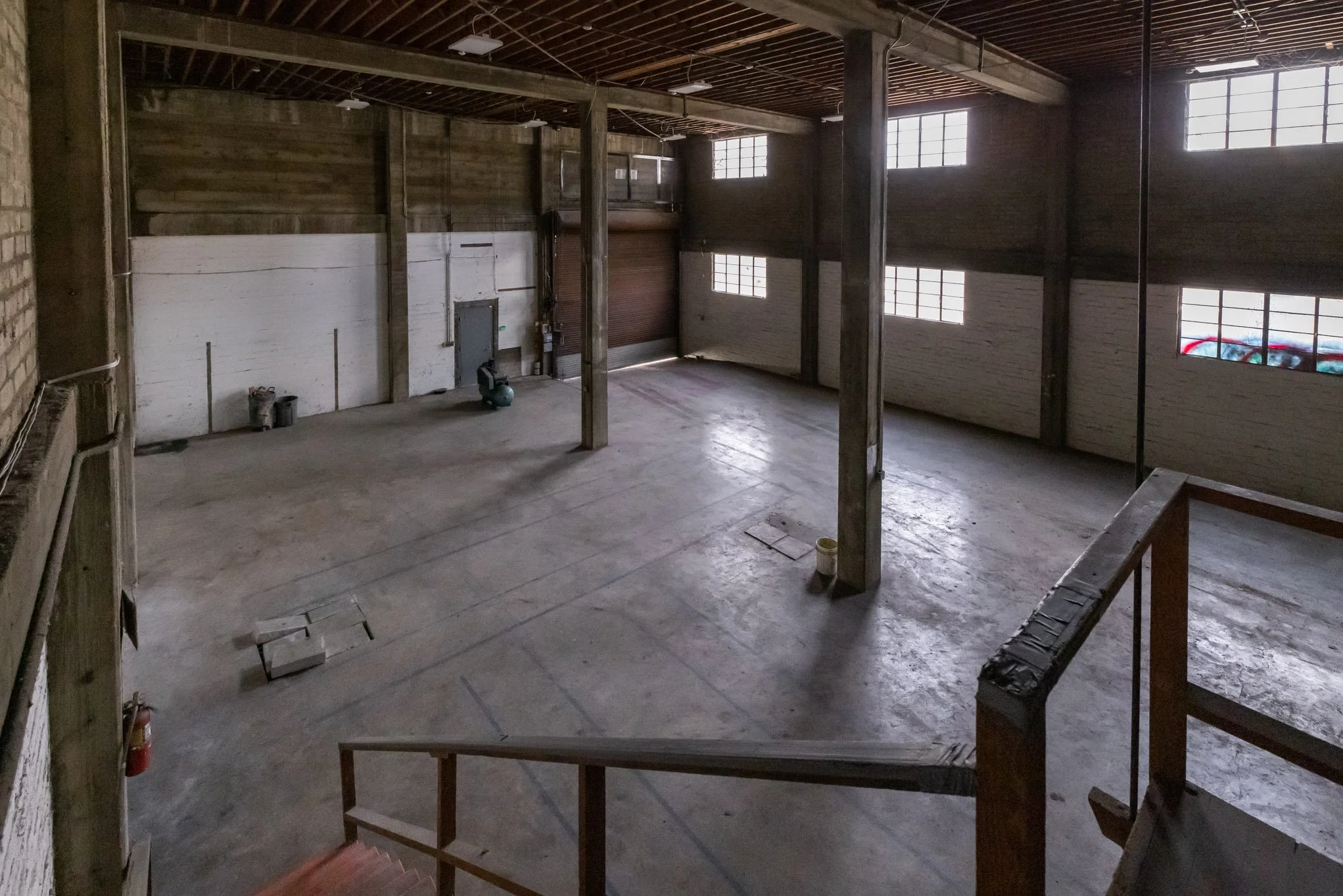 Empty industrial warehouse with high windows, concrete floor, wooden walls, and a staircase visible in the foreground.