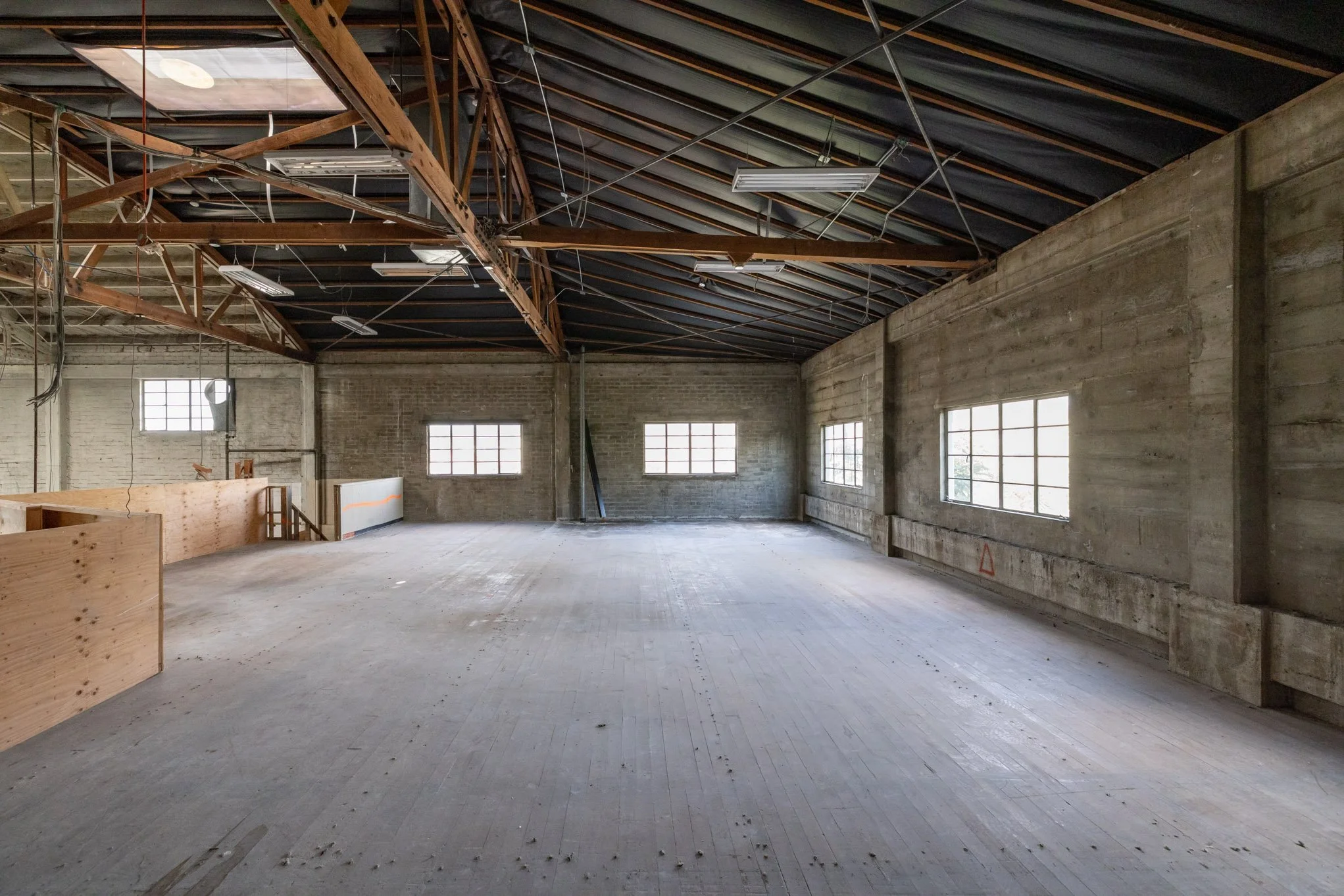 Empty industrial-style interior space with exposed wooden roof beams, large windows, and unfinished concrete flooring.