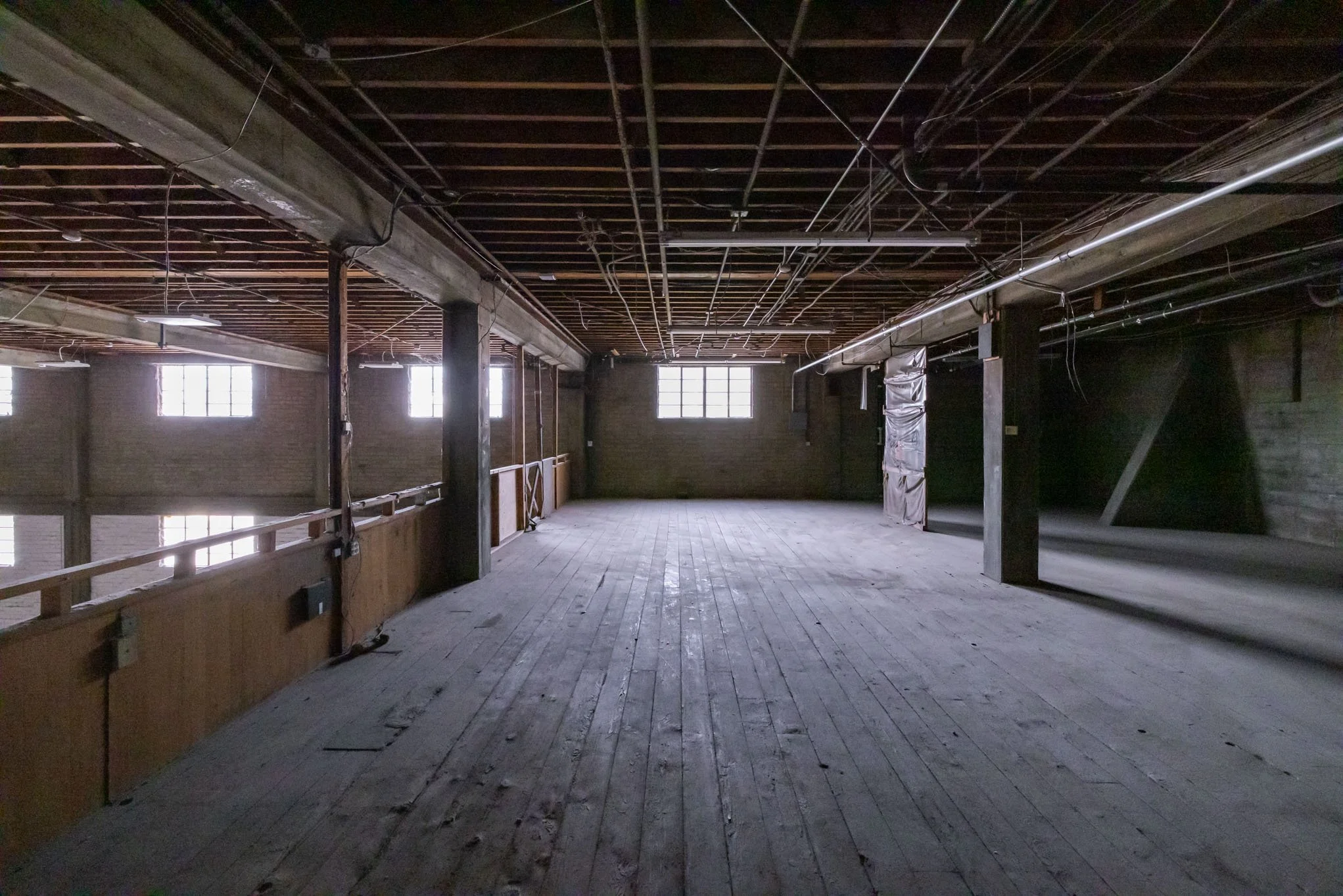 Empty, unfinished interior space with wooden floors, exposed ceiling beams, and brick walls with windows allowing natural light.