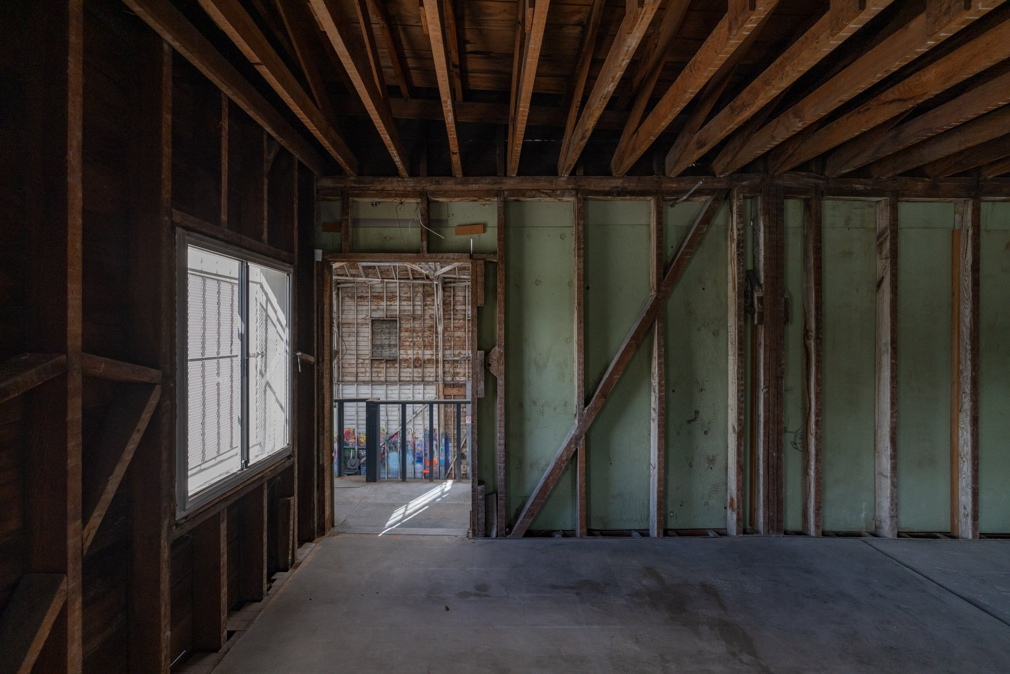 Interior of an unfinished room with exposed wooden framing, a small window on the left, and a door opening to an outside area with pallets and clutter.