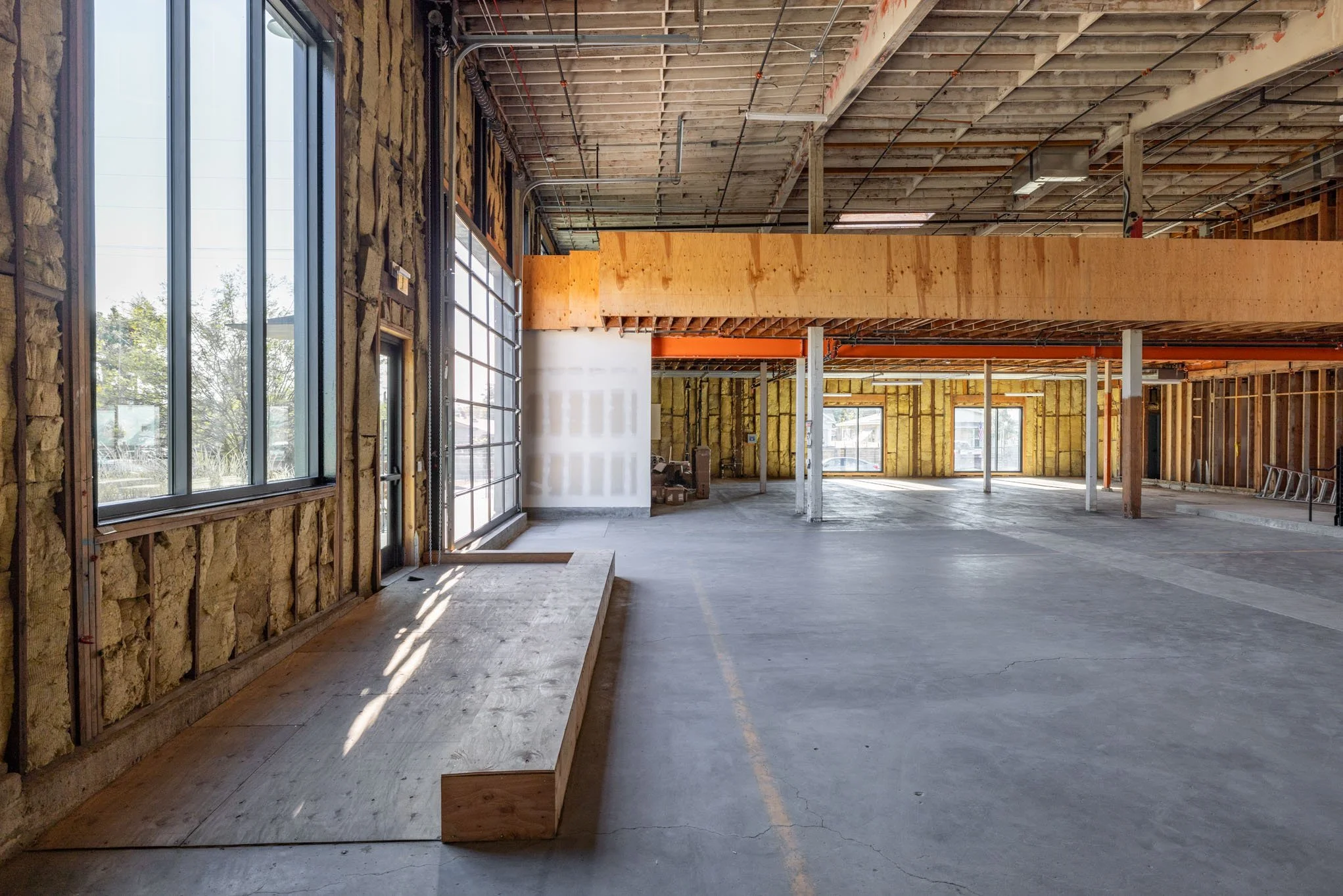 Interior of a building under construction with exposed walls, ceiling, and support beams, large windows, and an unfinished concrete floor.