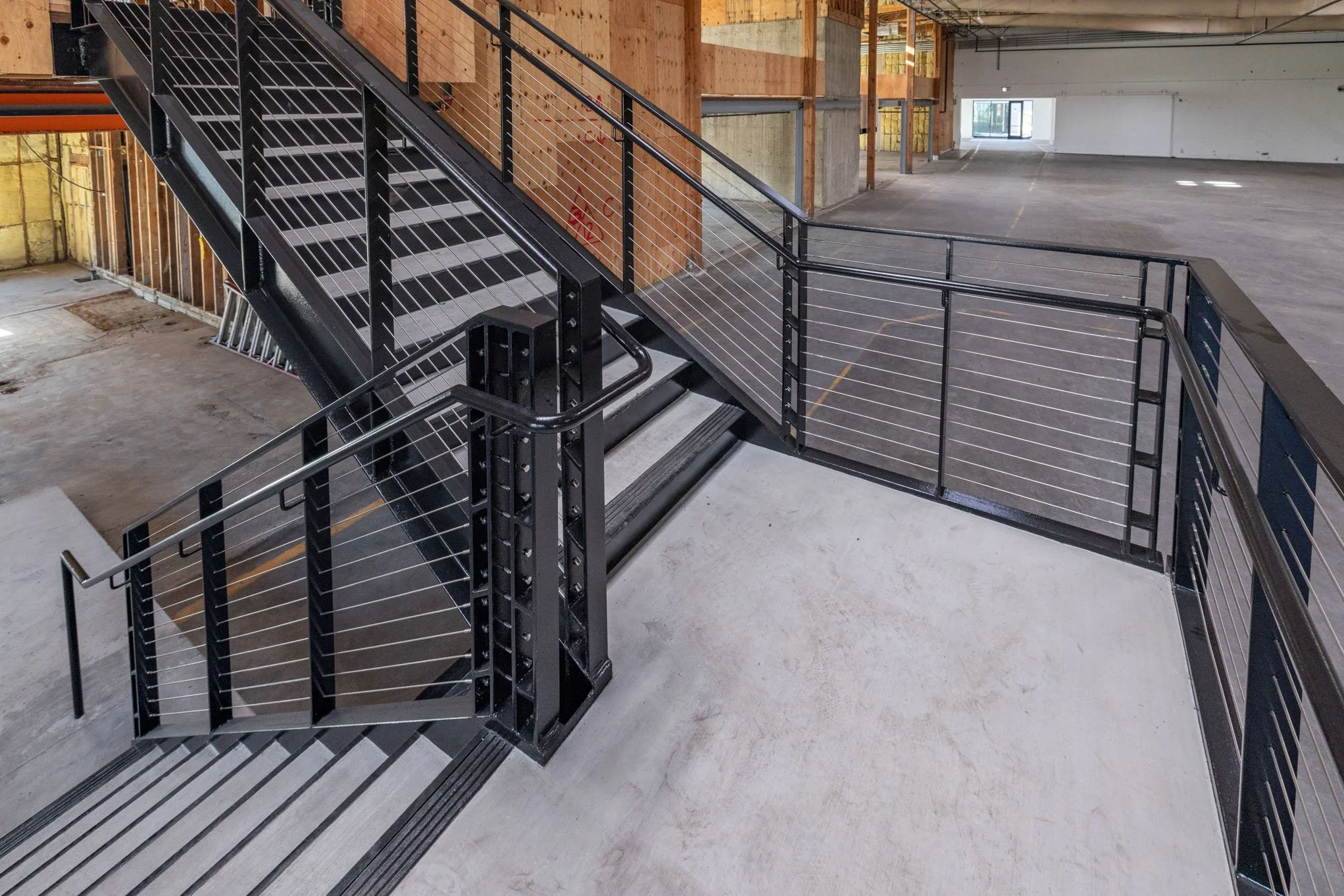 Interior view of a building under construction with a black metal staircase and safety railings, wooden framing on the walls, and an open space with a concrete floor.