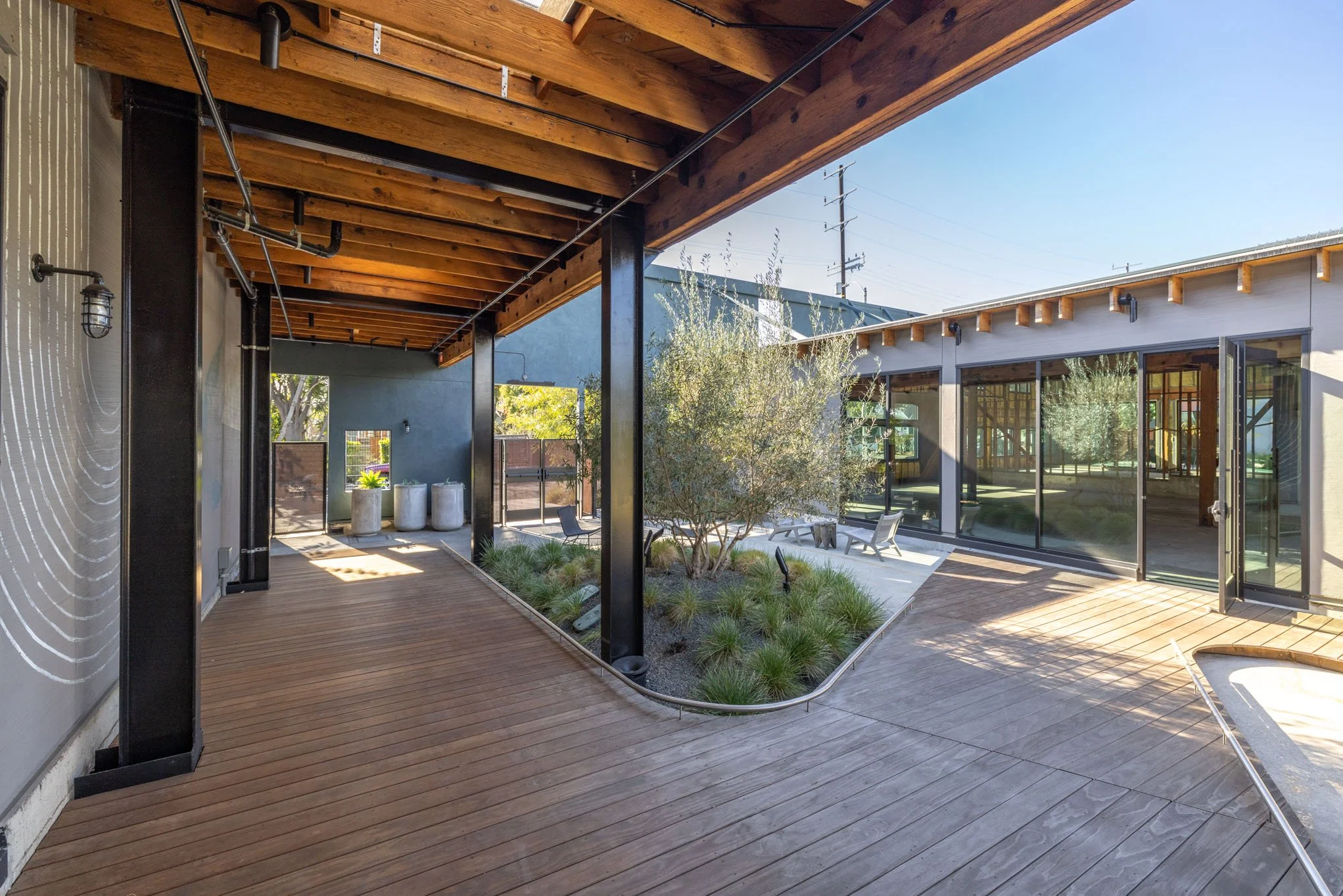 A modern outdoor patio with wooden decking, greenery in the center, and large glass sliding doors leading into a house.