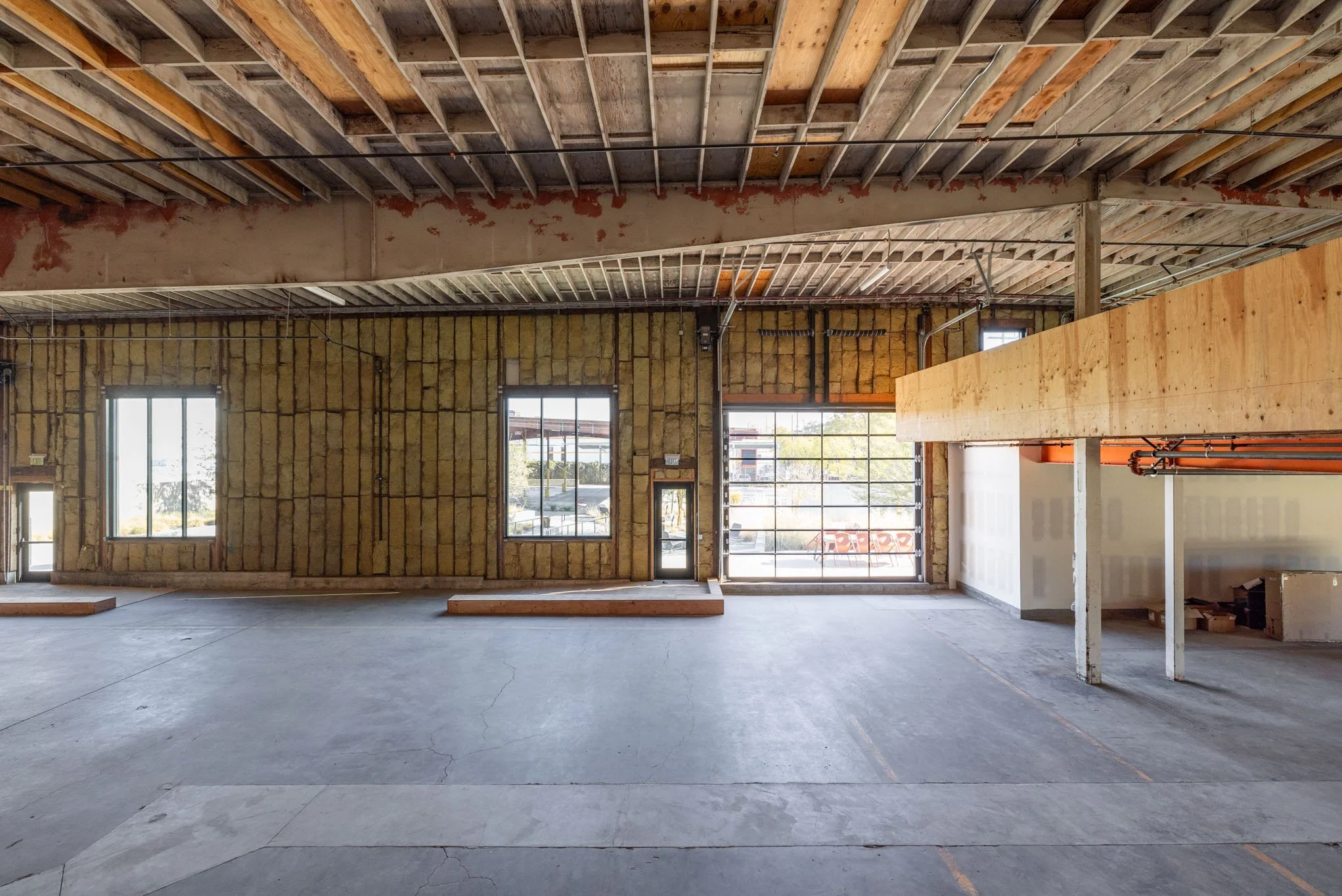 Empty retail or commercial space under construction with exposed ceiling beams, unfinished walls, large windows, and a glass garage door.