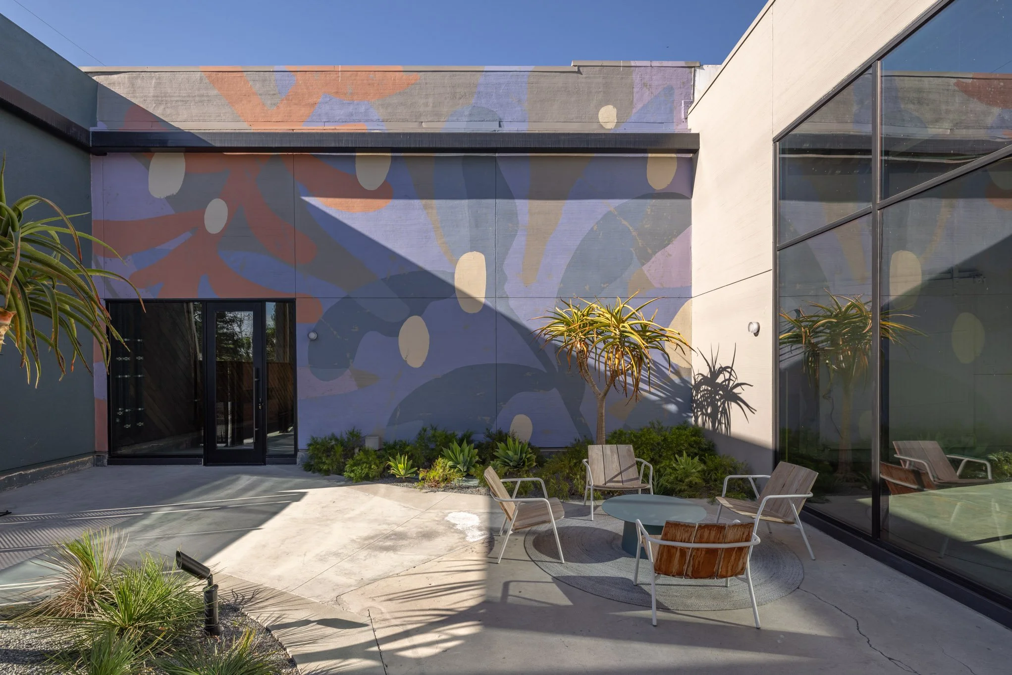 Outdoor patio with modern furniture, small round table, four chairs, and plants, including a large mural with abstract shapes on the building wall, glass windows reflecting the sky.