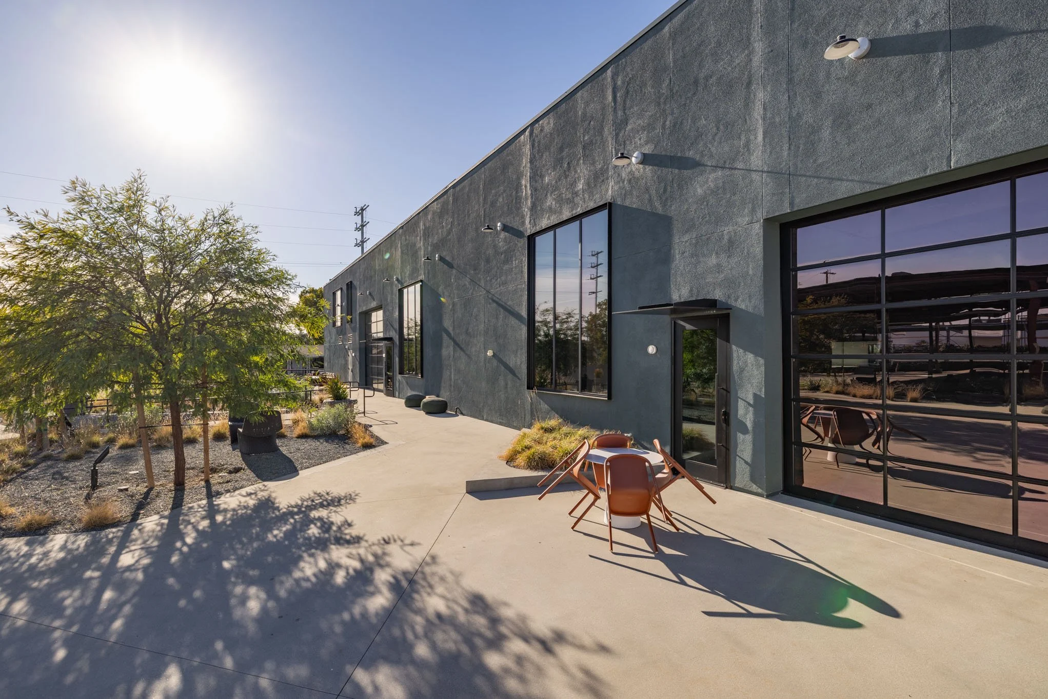 Outdoor patio area with tan chairs, potted plants, a tree, and a gray modern building with large windows, under clear sunny sky.