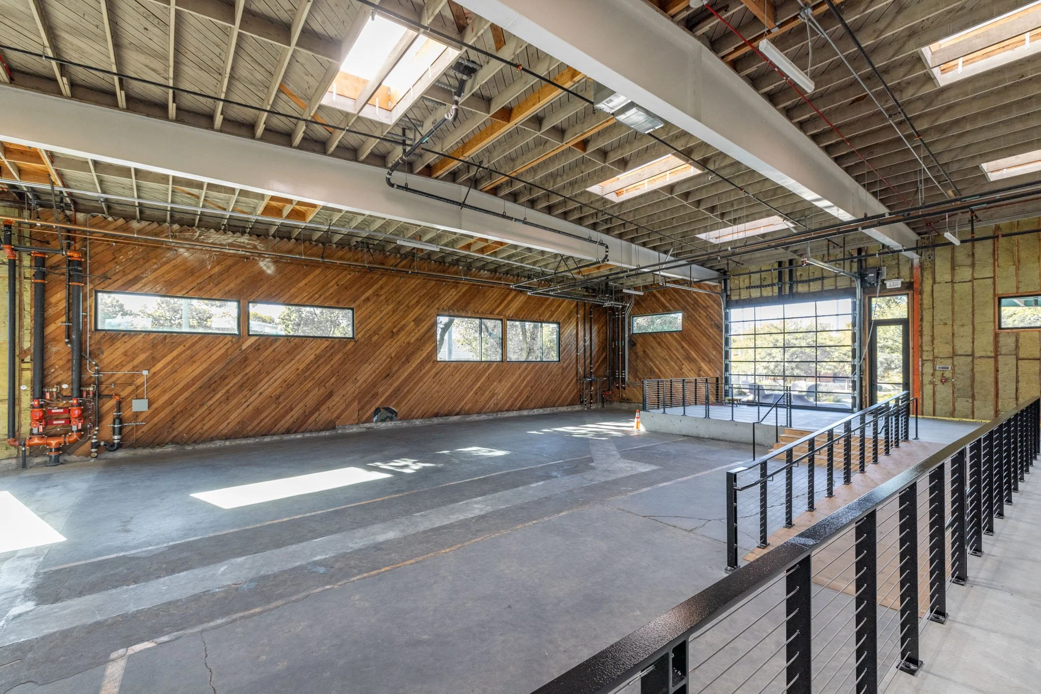 Empty indoor space under construction with partly finished wooden wall, large windows, and a glass garage door, featuring exposed ceiling beams and skylights.