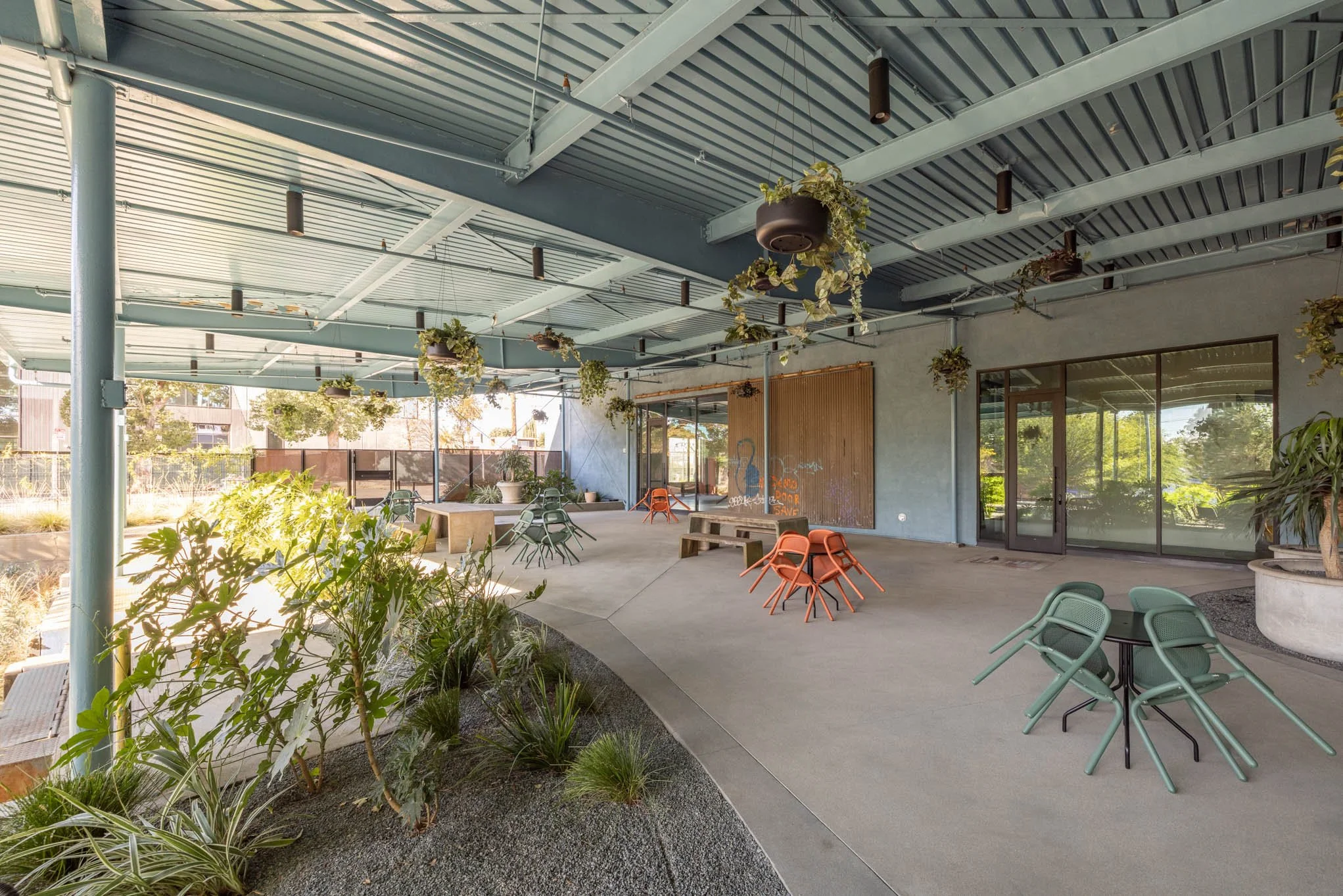 Open-air patio with hanging plants, green and orange chairs, potted plants, and large glass windows.