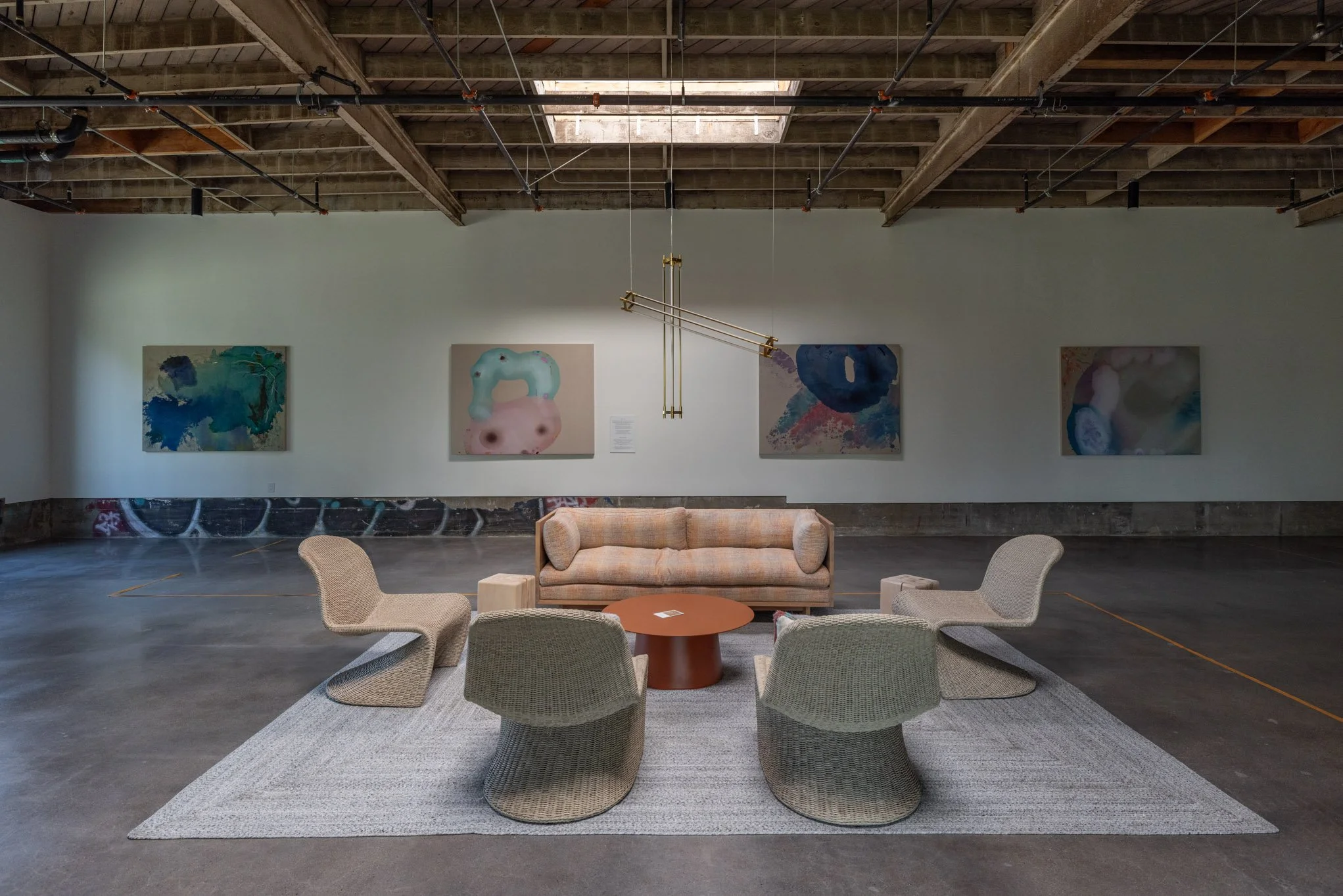 An art gallery with four large abstract paintings on the wall, a beige sofa, four modern chairs, a round wooden table, and a gray rug on a polished concrete floor.