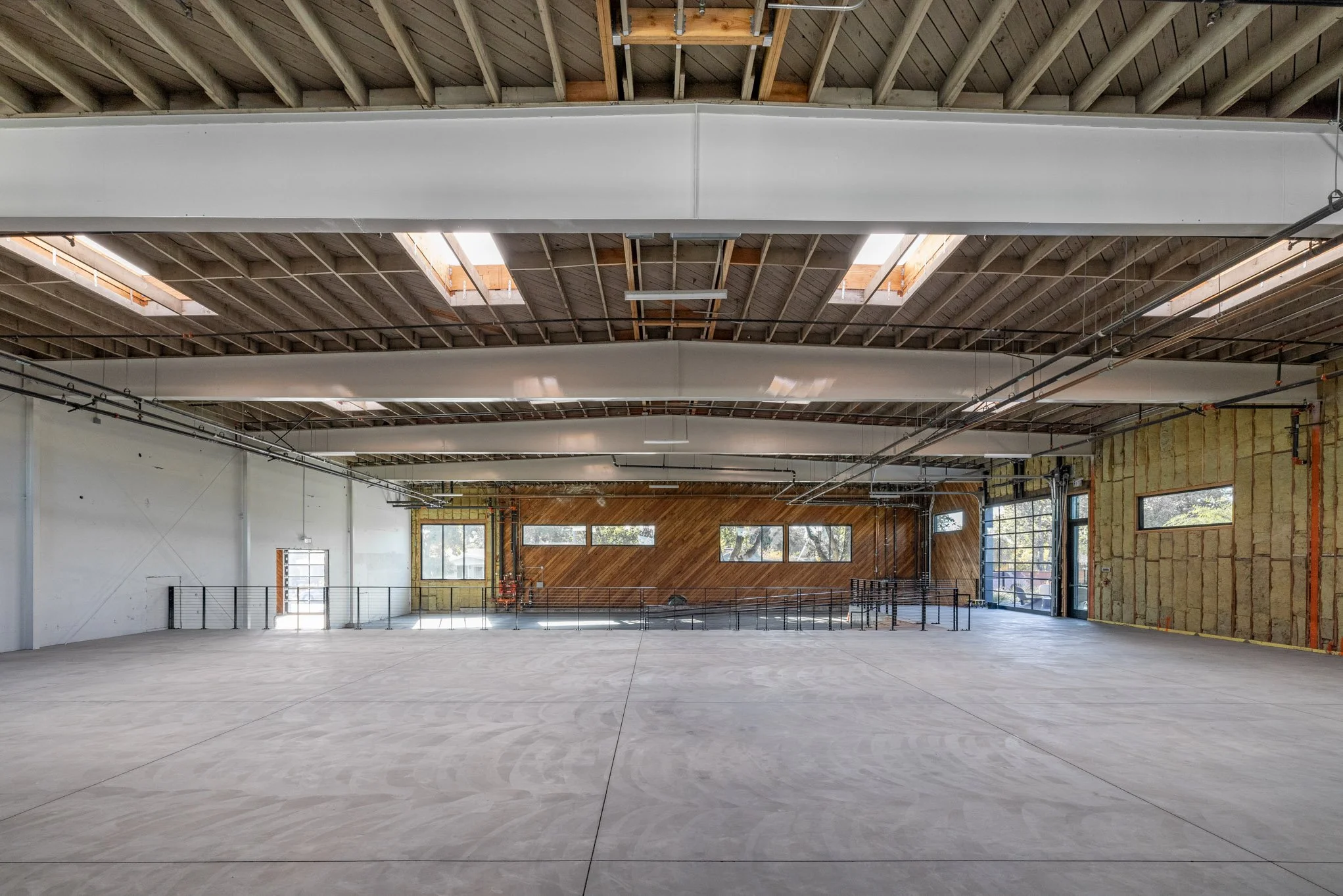 An empty indoor space under construction with exposed wooden ceiling beams, partial drywall, and large windows, featuring a raised wooden wall and a railing, likely a gym or community hall.