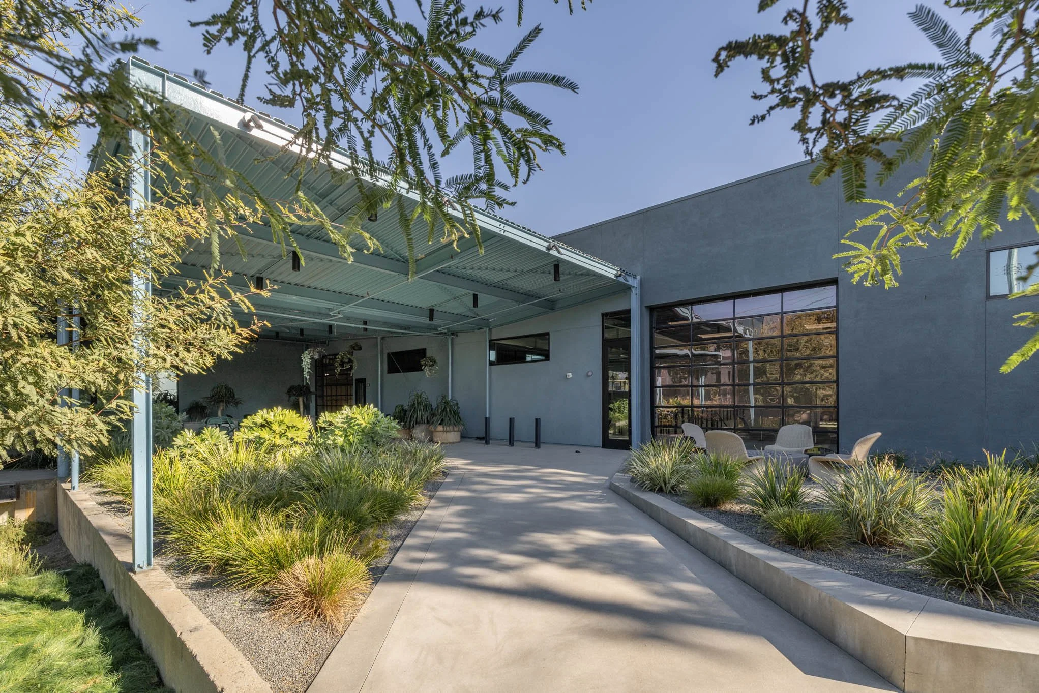 Modern building with large glass windows and a patio area, surrounded by landscaped plants and trees, with a blue sky overhead.