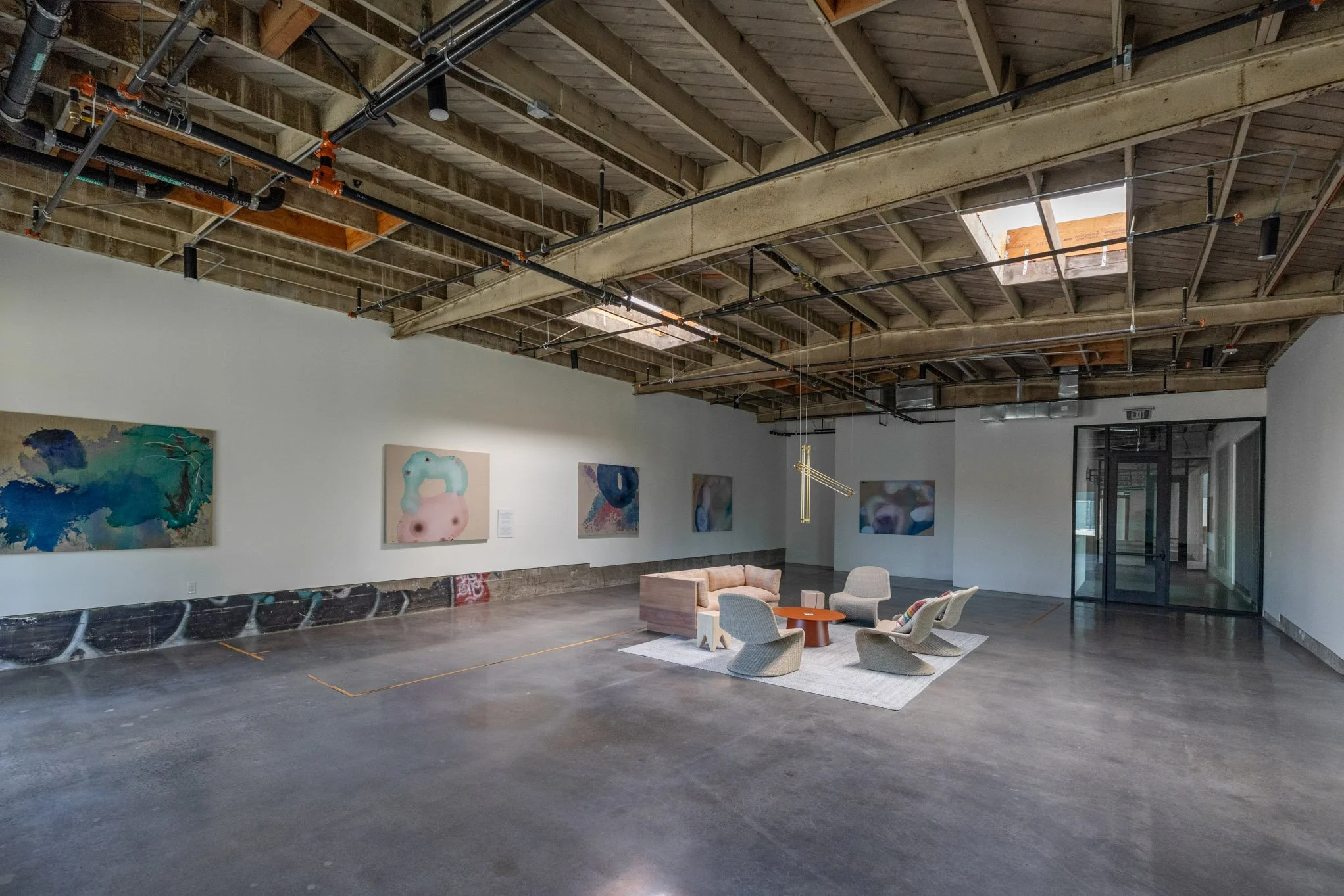 Interior view of an art gallery with abstract paintings on white walls, a seating area with four modern chairs, a small table, and a beige sofa on a white rug, all under an unfinished wooden ceiling with skylights and exposed pipes.
