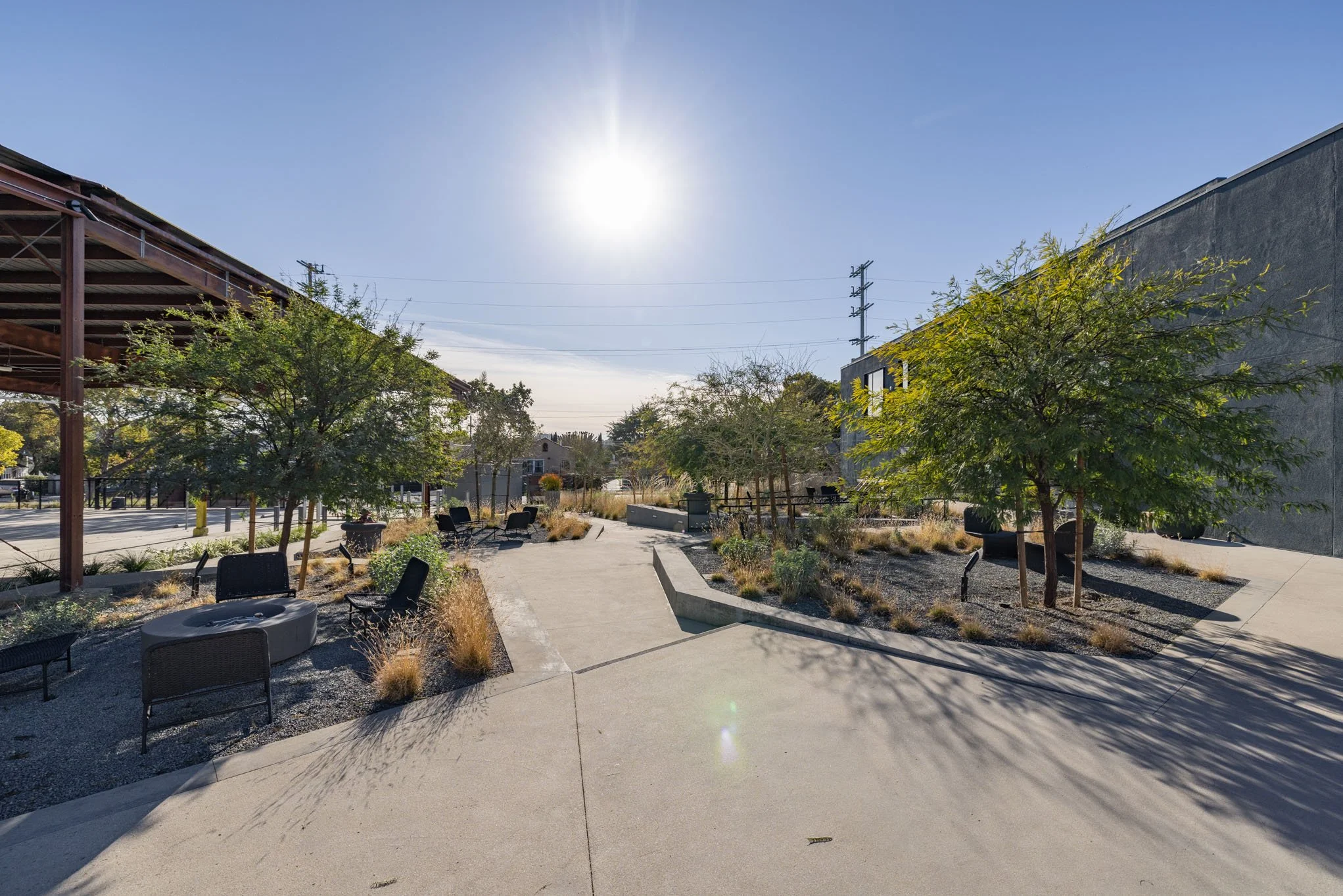 A sunny outdoor park area with trees, benches, and a paved walking path, with a building on the right and a covered pavilion on the left.