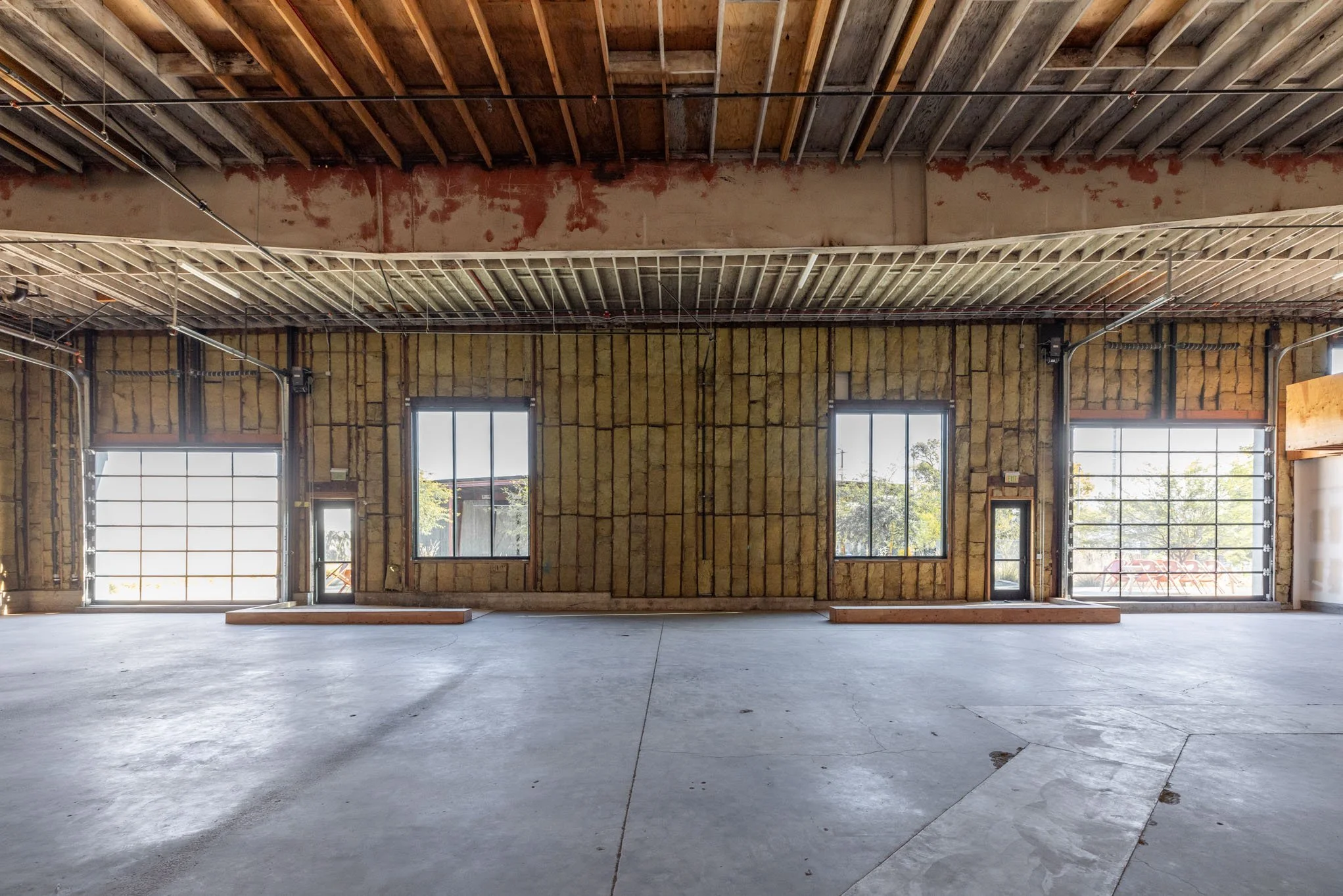 Empty interior of a building under construction with exposed insulation and framing, large windows, and garage-style doors.