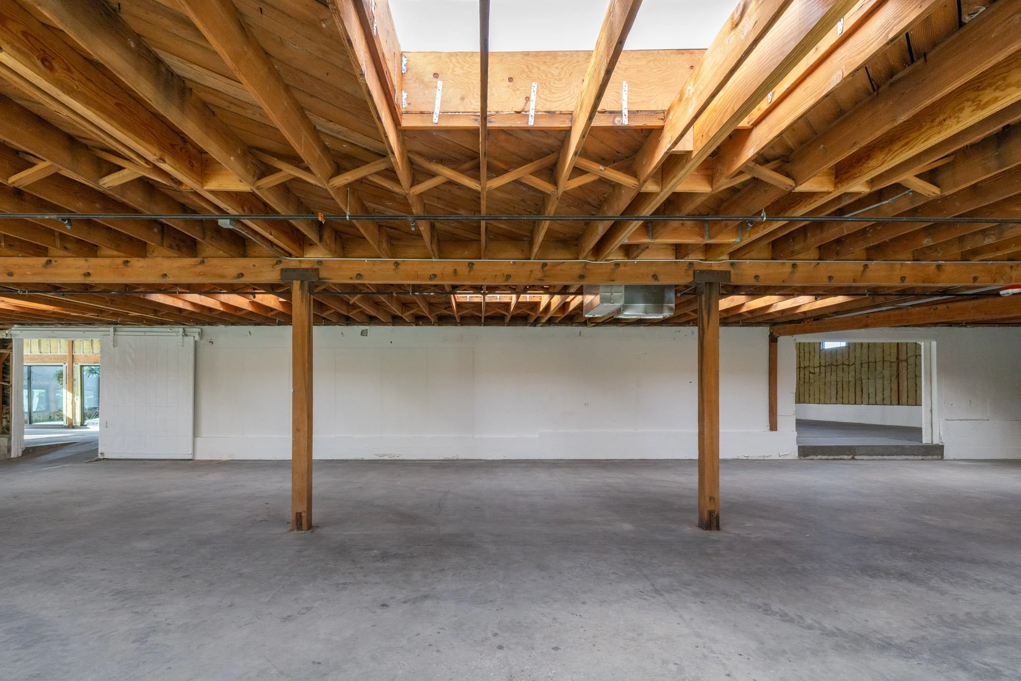 Empty loft space with exposed wooden ceiling beams and support columns, concrete floor, and unfinished walls, with a small opening on the right side and sliding door on the left.