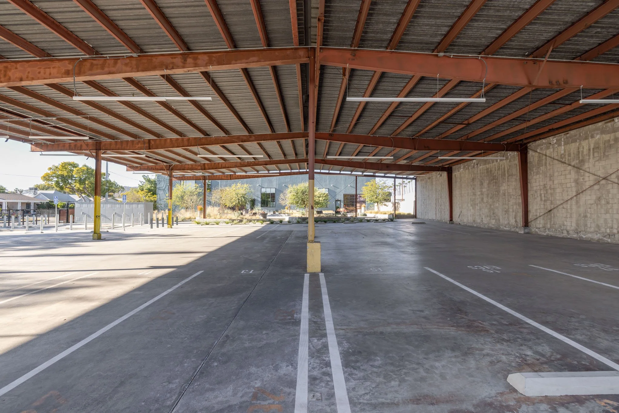 Empty parking lot with marked parking spaces under a metal roof structure with support beams, with some trees and a building visible in the background.