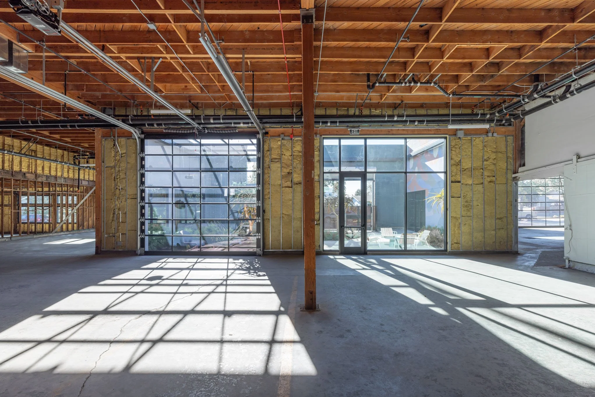 Empty indoor space with unfinished walls, large glass garage door, glass entrance door, exposed ceiling beams, and sunlight streaming in, casting shadows on the concrete floor.