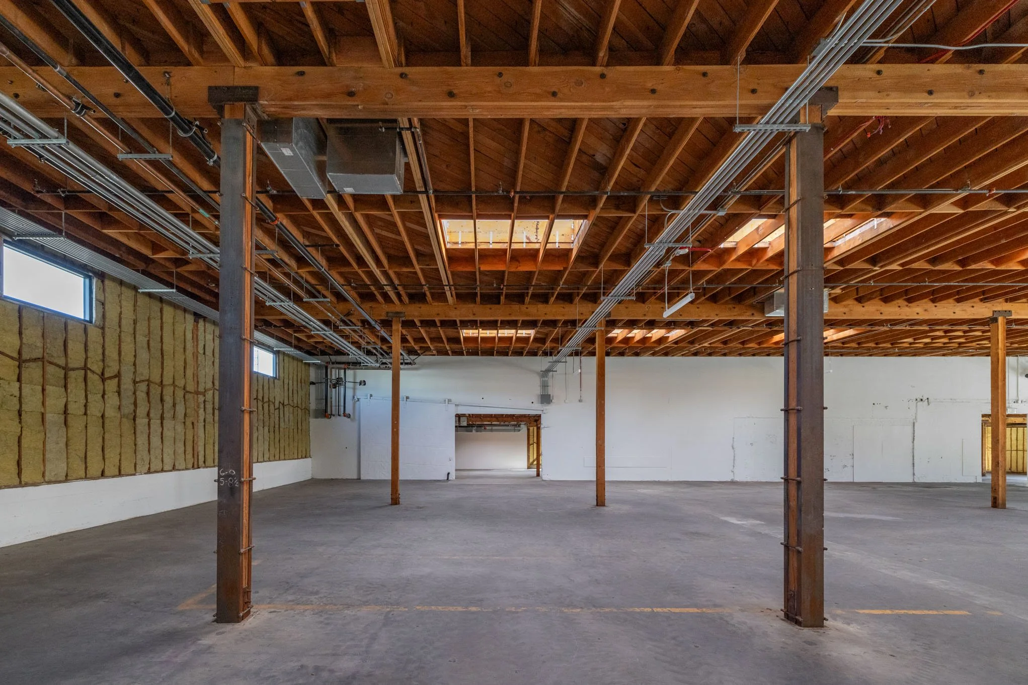 Empty indoor warehouse or industrial space with exposed wooden ceiling beams, metal electrical conduits, and support columns, with small windows on the left wall.