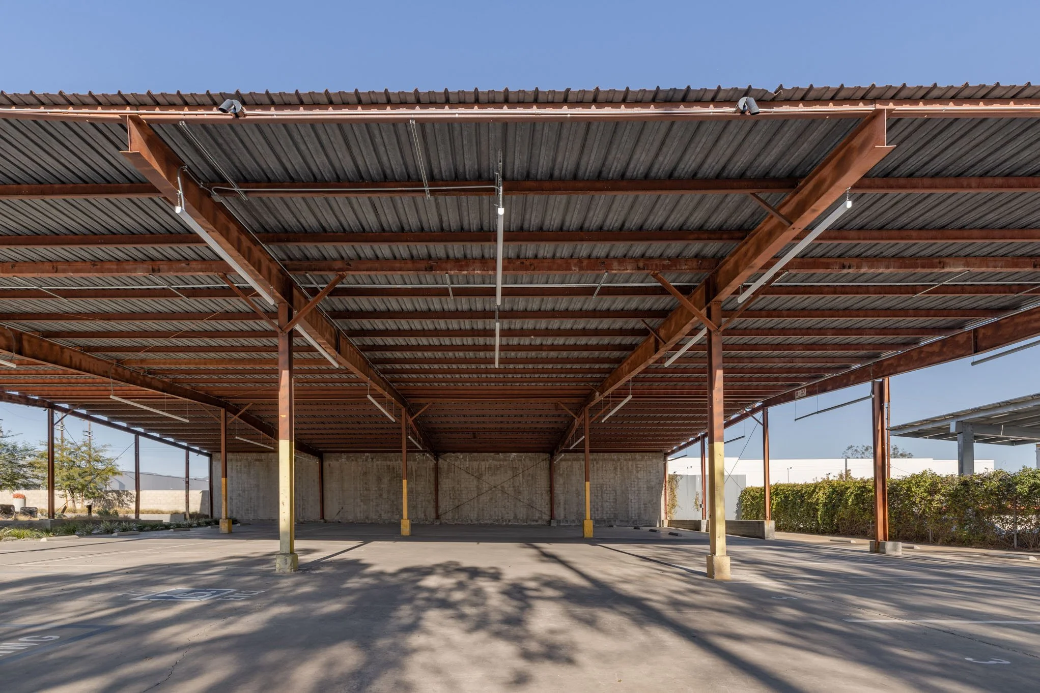 Empty outdoor parking lot with a metal roof structure, shadows cast by nearby trees, and surrounding bushes.