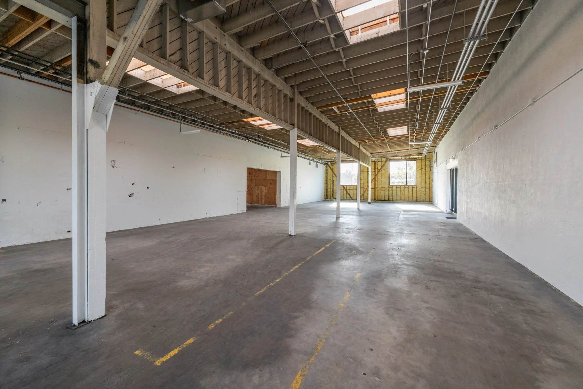 Empty, unfinished interior space with concrete floors, white walls, exposed ceiling beams, and windows at the far end, in a building under construction or renovation.