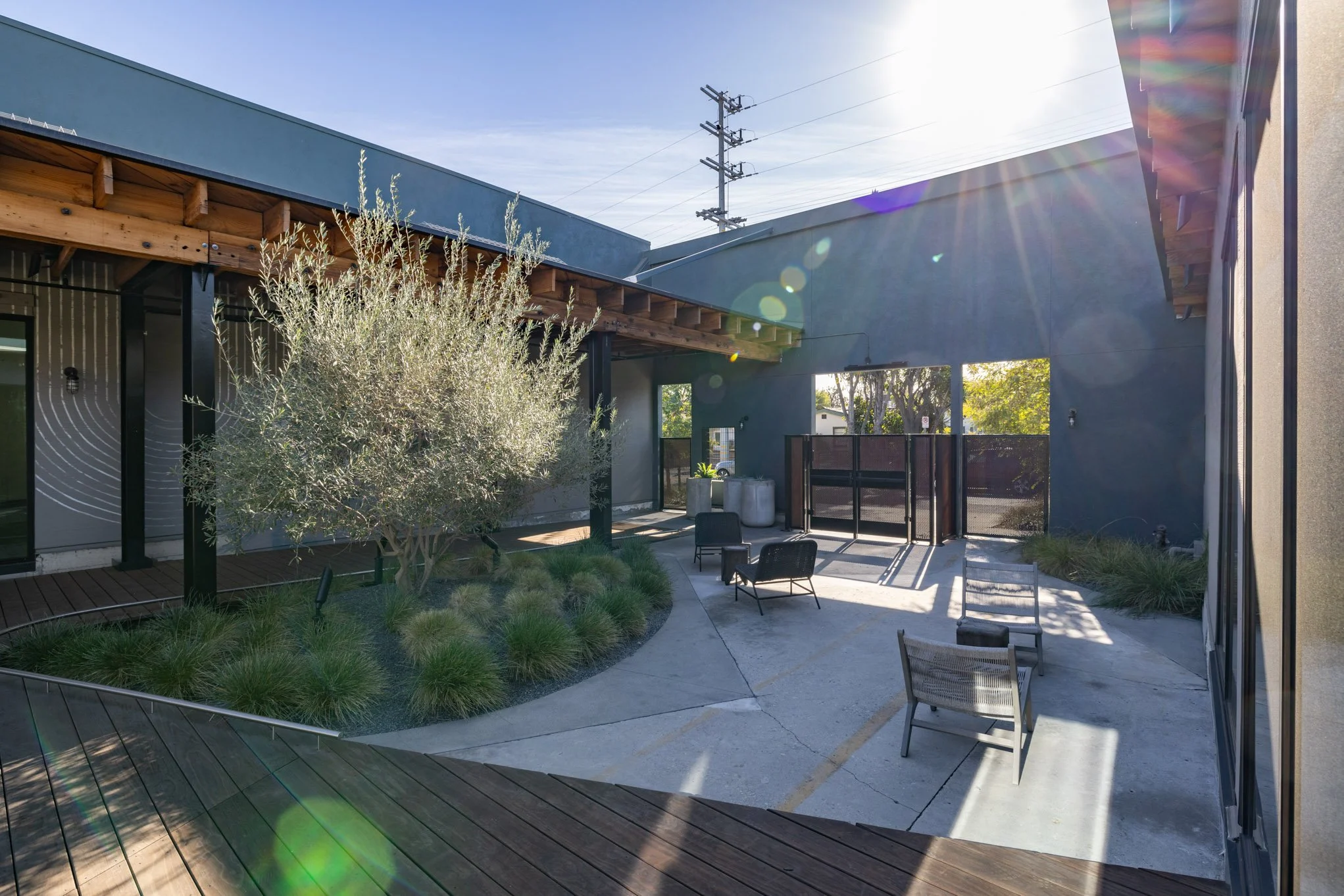 Outdoor patio with modern furniture, plants, and a concrete ground, surrounded by buildings with a wooden overhead structure and clear sky with sunlight.