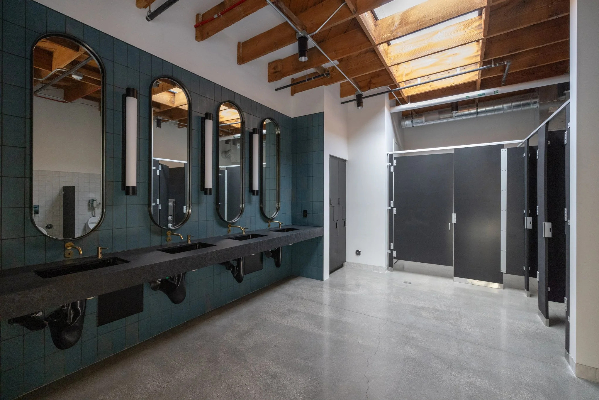 Public bathroom with three sinks, tall oval mirrors, black fixtures, and gray tiles, with dark stalls and wooden ceiling beams, illuminated by natural light from a skylight.