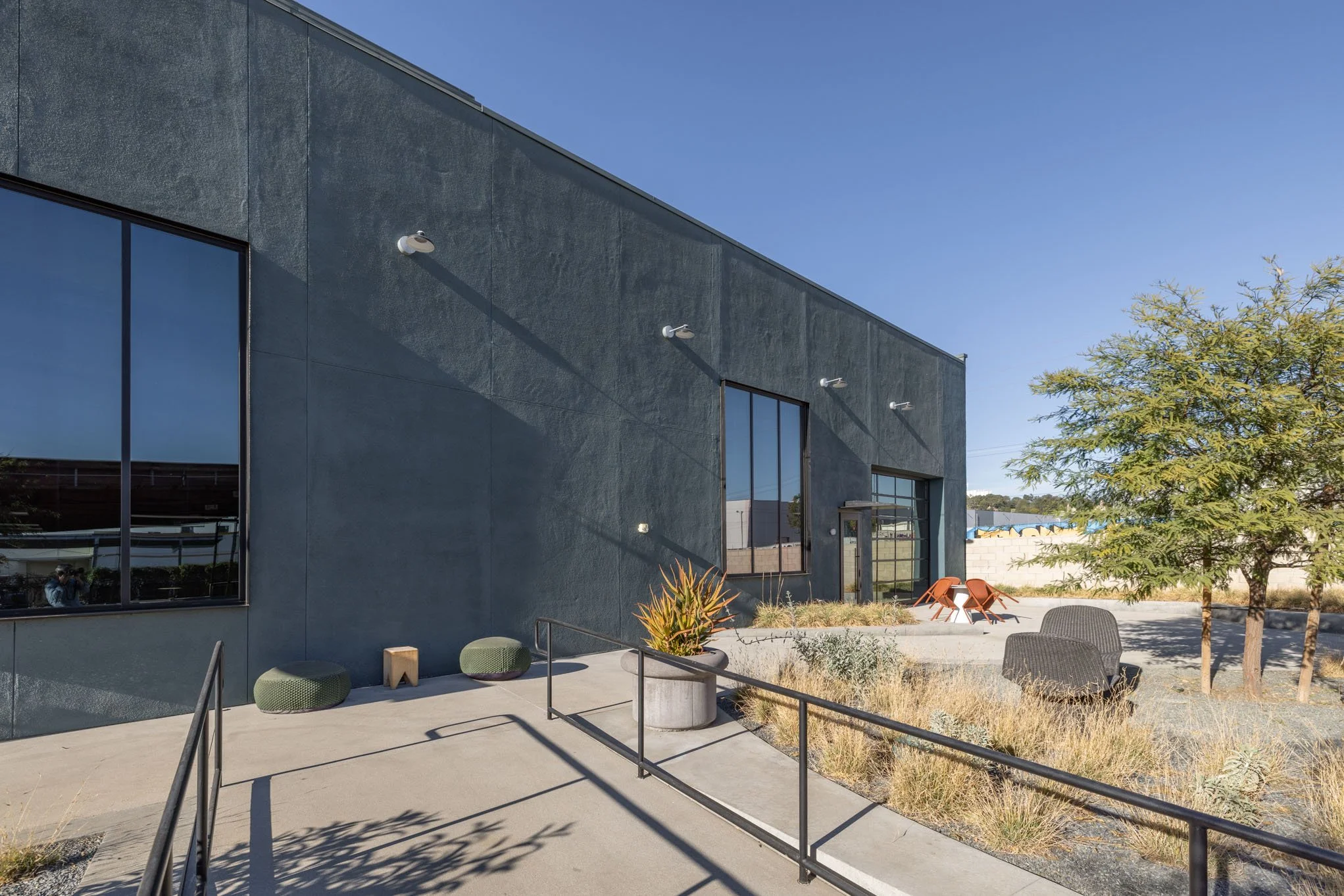 Exterior view of a modern building with large windows, outdoor seating, plants, and a tree in a sunny area.