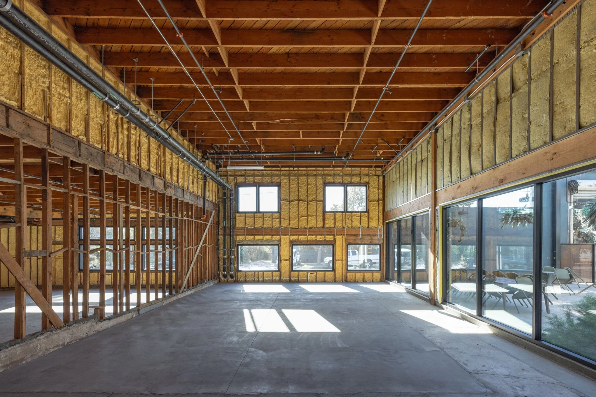 An interior view of a building under construction with exposed wooden ceiling beams, yellow insulation, unfinished walls, and large glass windows, allowing sunlight to fill the space.