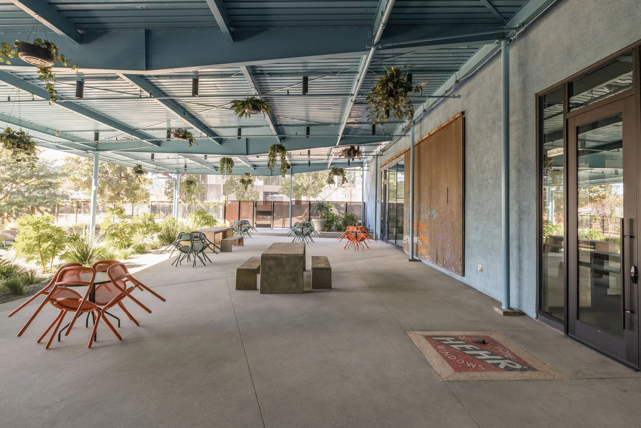 Empty outdoor patio area with modern chairs, benches, hanging plants, and an open sky roof, adjacent to a building with large glass doors and a concrete wall.