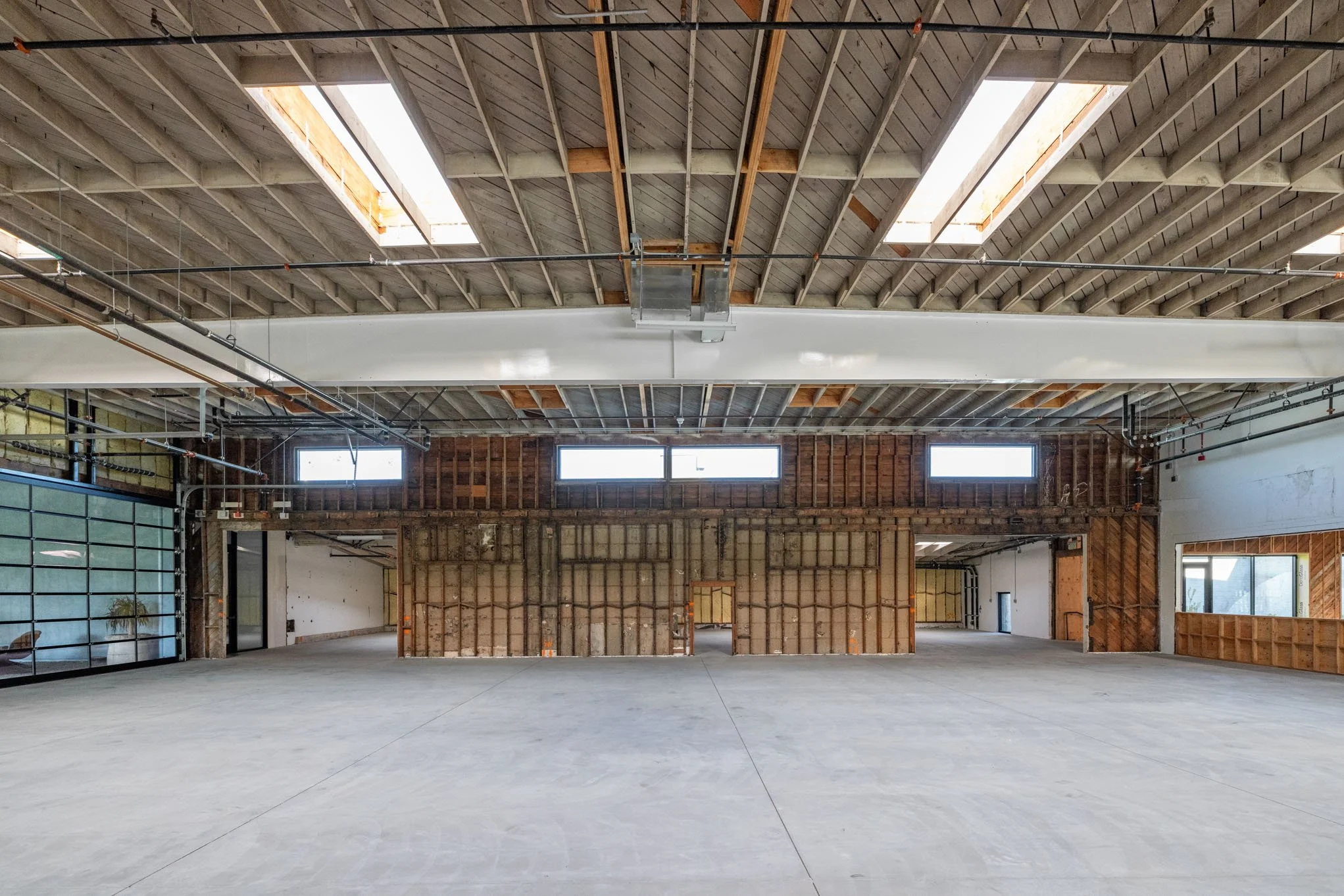 Large industrial space under construction with exposed wooden ceiling and walls, concrete floor, and several windows providing natural light.