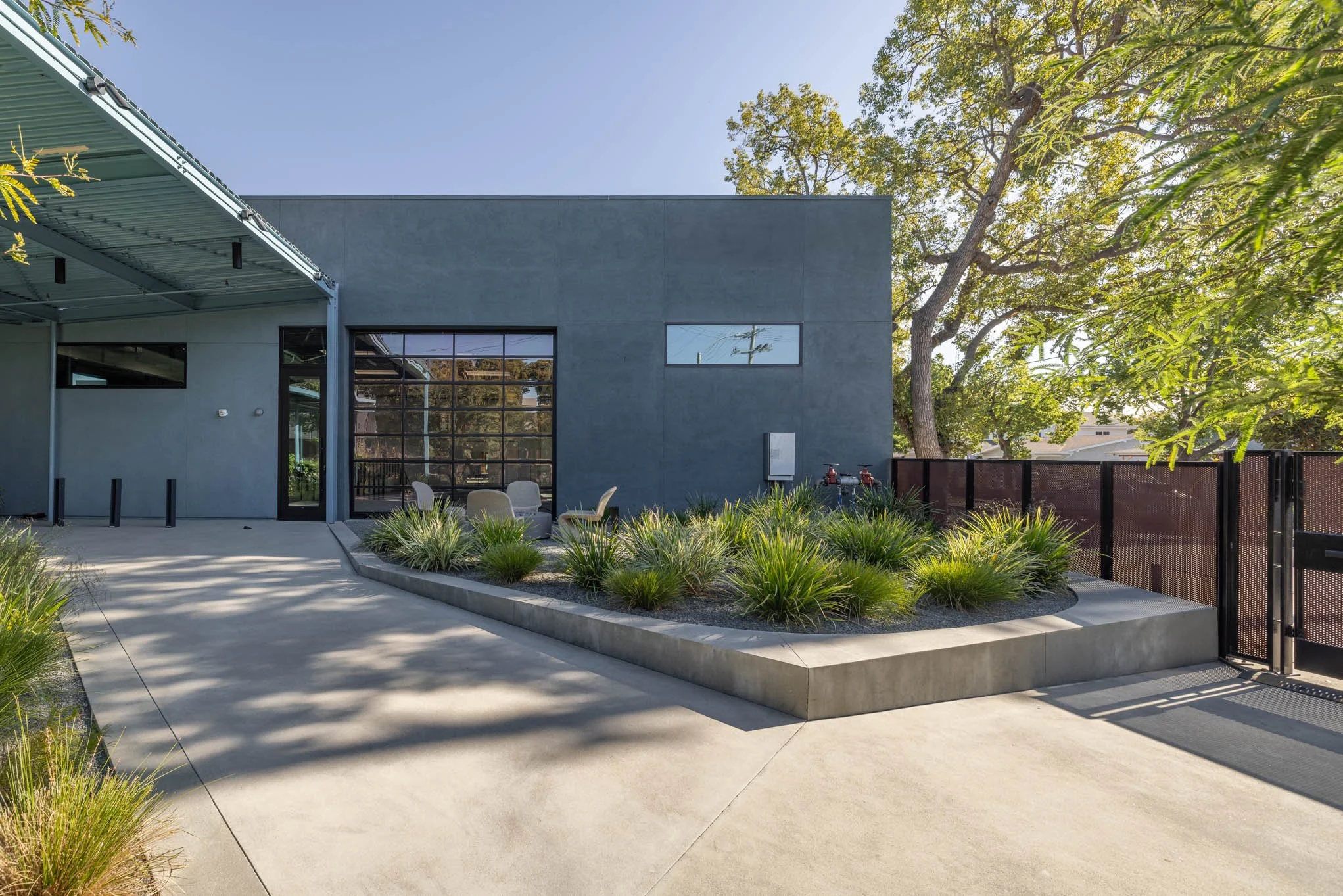 Modern building exterior with grey walls, large glass windows, and a landscaped area with plants and seating. Contains a short pathway, trees, and a black fence.